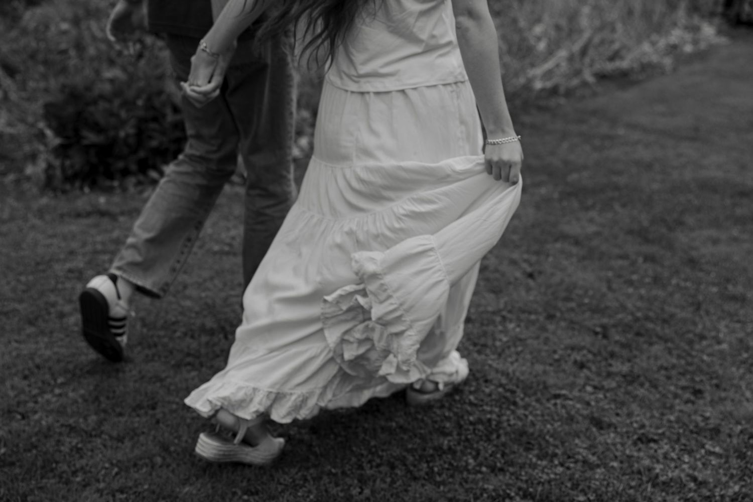 Black and white photo of a couple walking on grass, woman in flowing white wedding dress with ruffled hem.