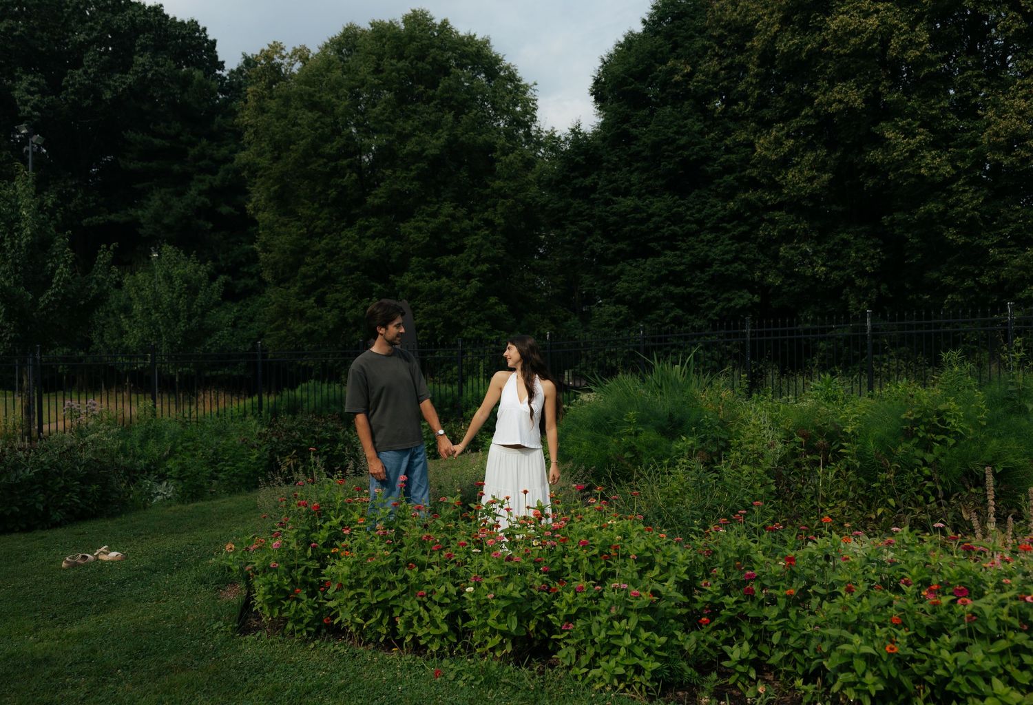 A couple holds hands while walking through a colorful flower garden surrounded by tall green trees.