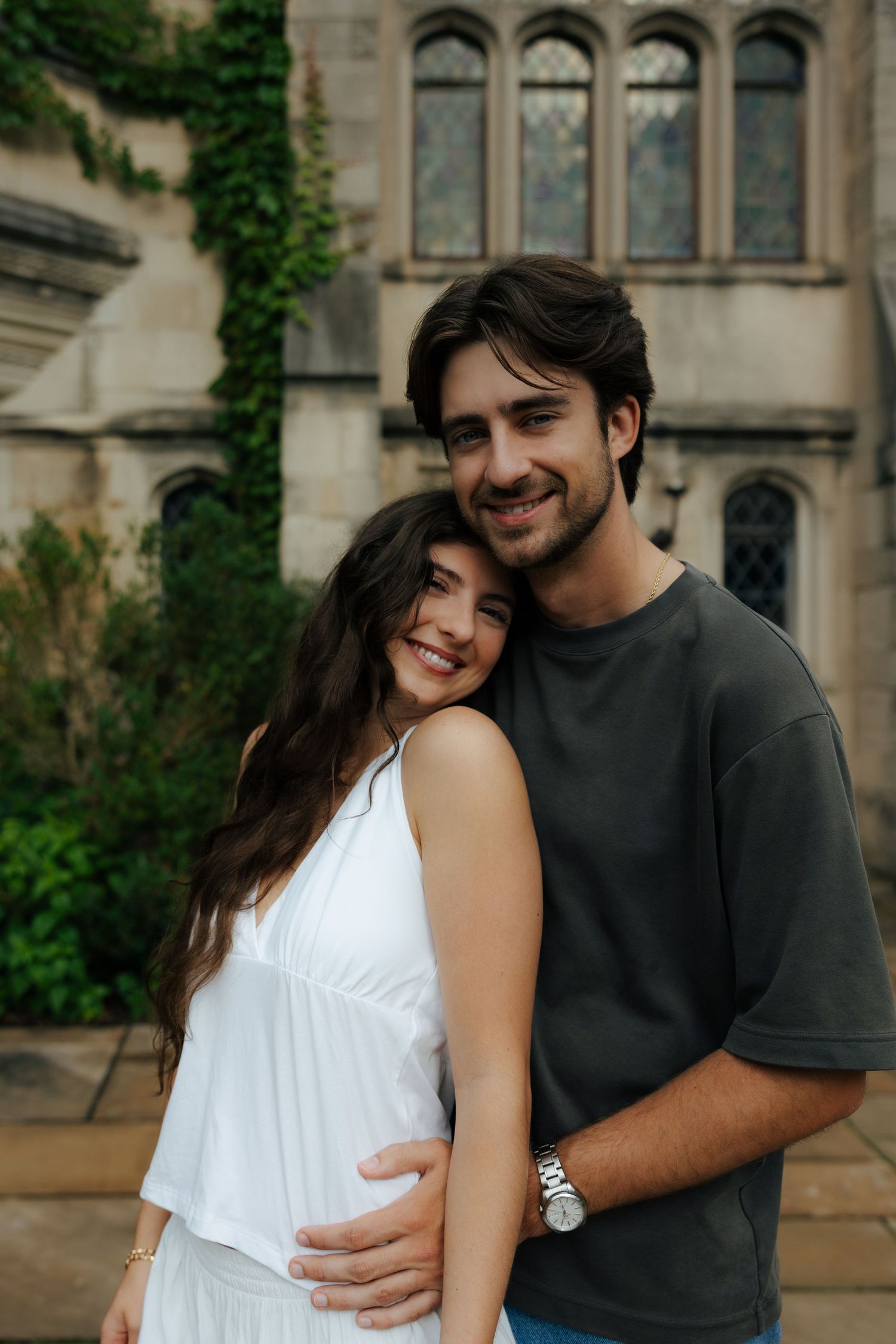 A smiling couple embraces in front of a gothic stone building covered in lush green ivy.