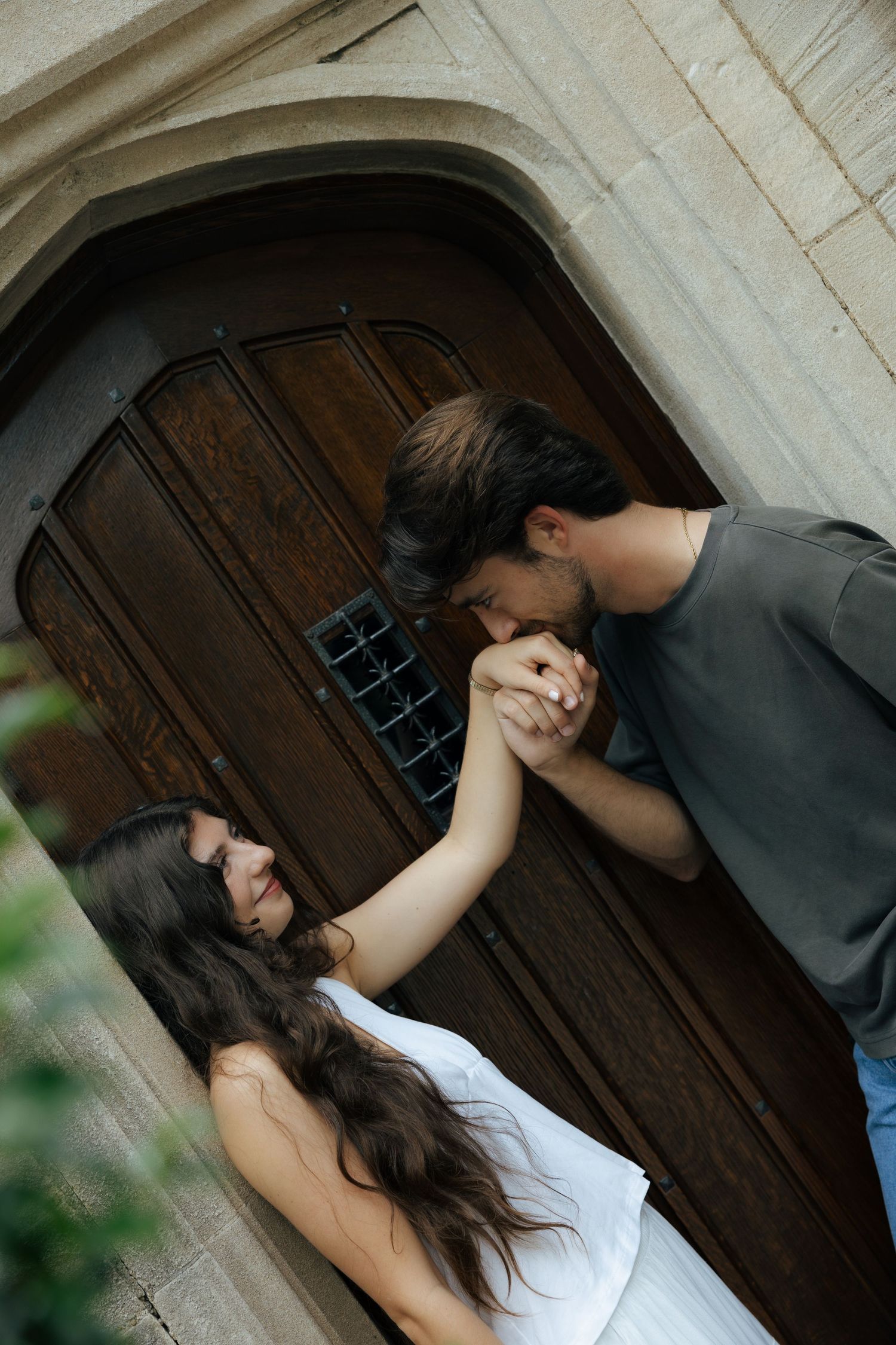 Man kissing woman's hand in front of an ornate wooden arched door at a stone building.