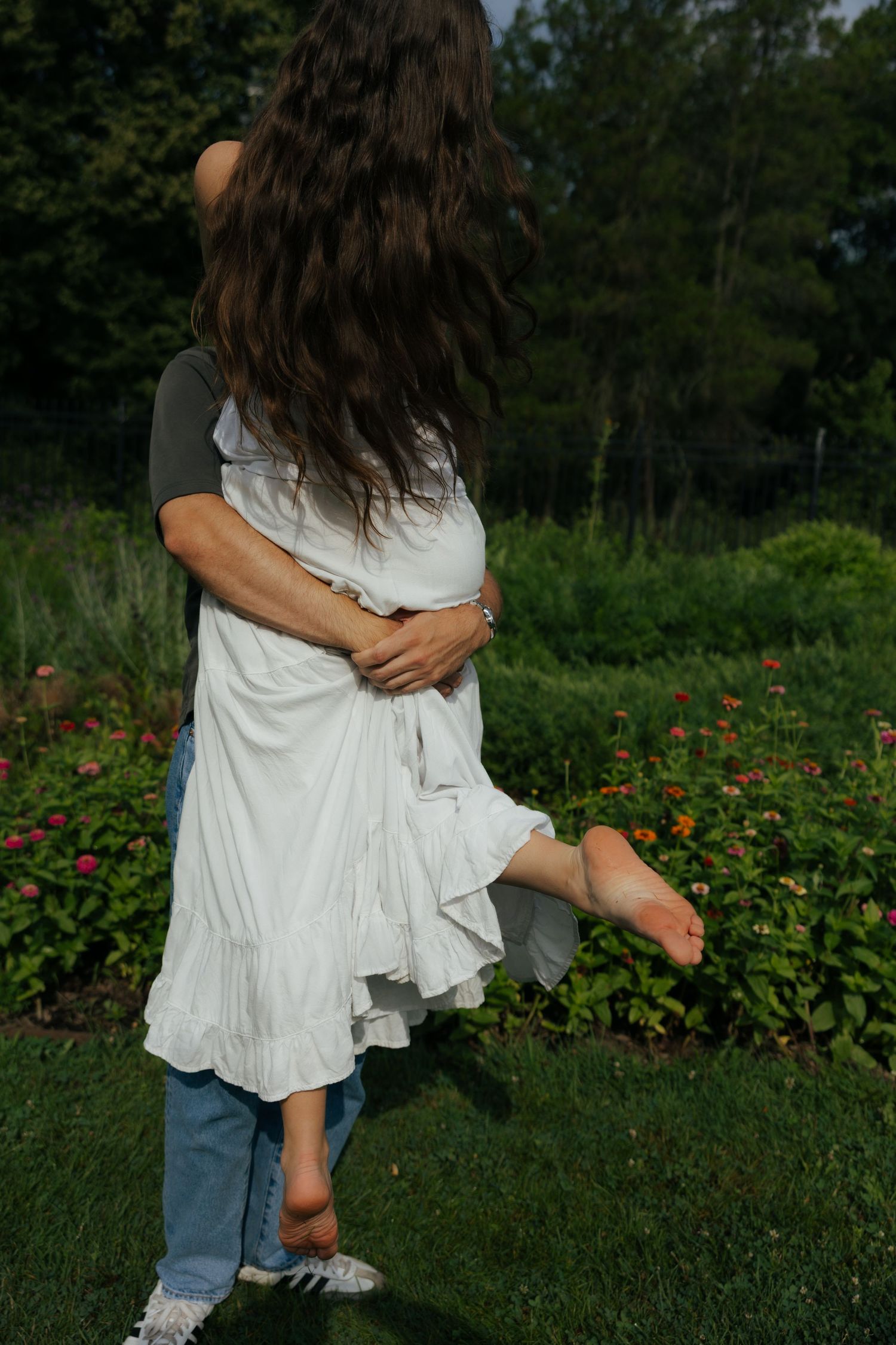 Man embracing woman in white dress from behind in a sunny garden with colorful flowers.