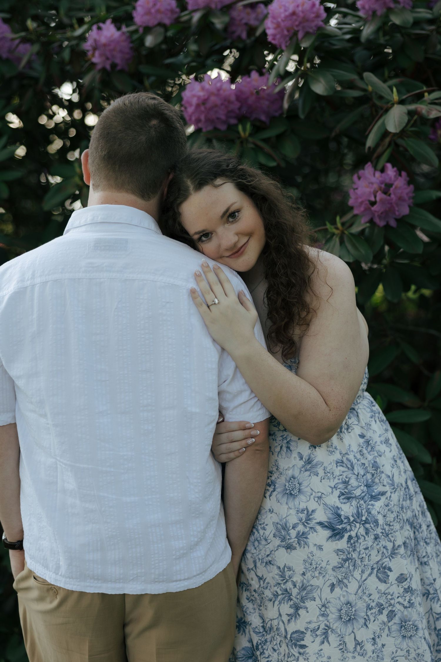 Woman in floral dress smiling at camera while embracing man from behind near purple flowering bushes.