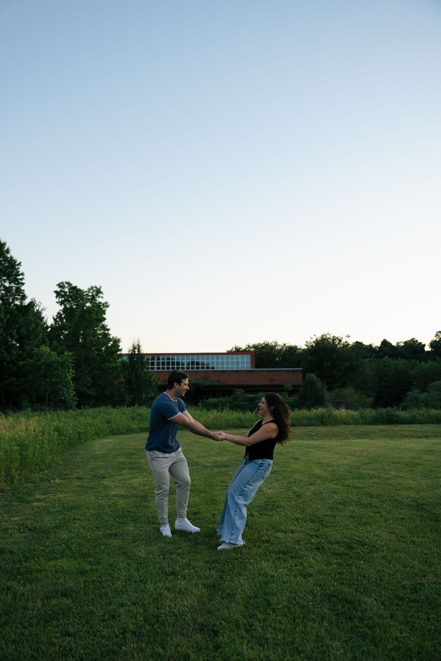 An engaged couple holds hands and spins together on a green lawn during a golden hour photo session.