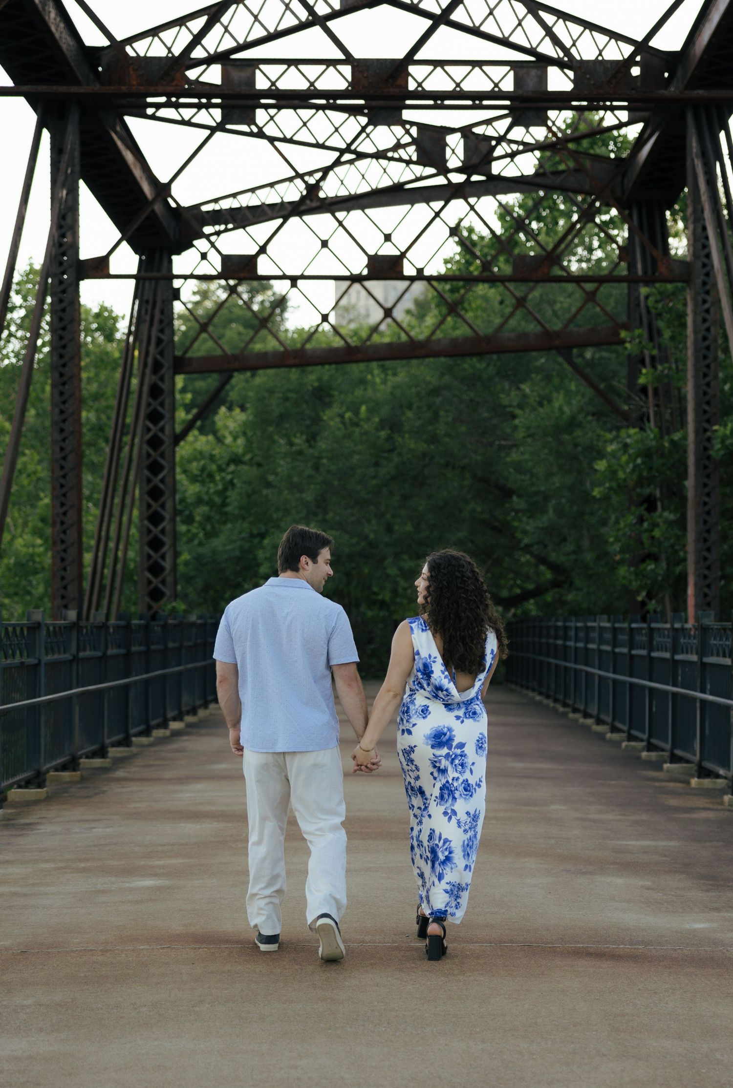 A couple walks hand in hand away from the camera down a scenic iron truss bridge pathway.