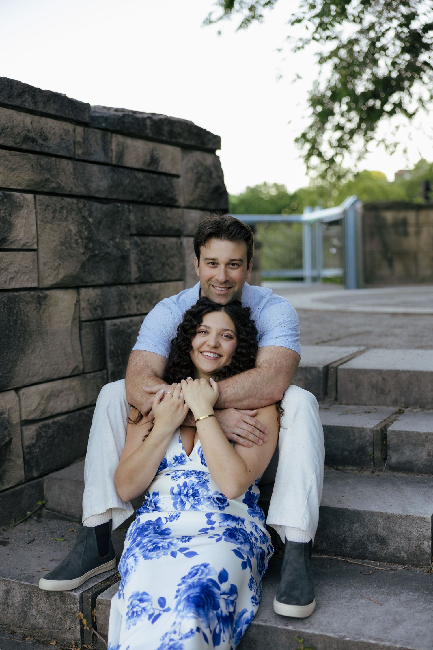 Happy couple sitting on stone steps outdoors, man hugging woman from behind, both smiling.