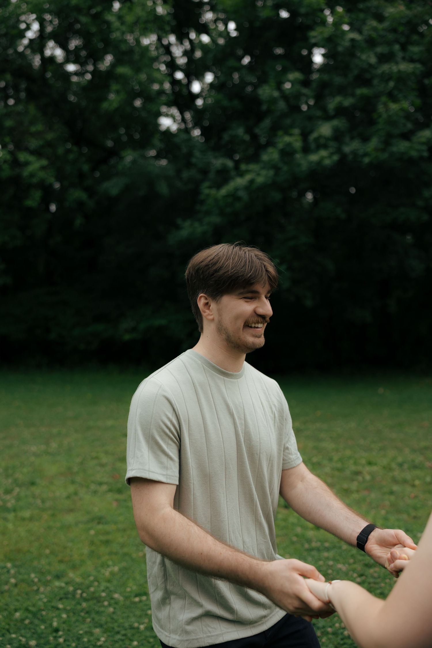 Smiling young man in a light gray t-shirt holding hands with someone in a lush green park setting.