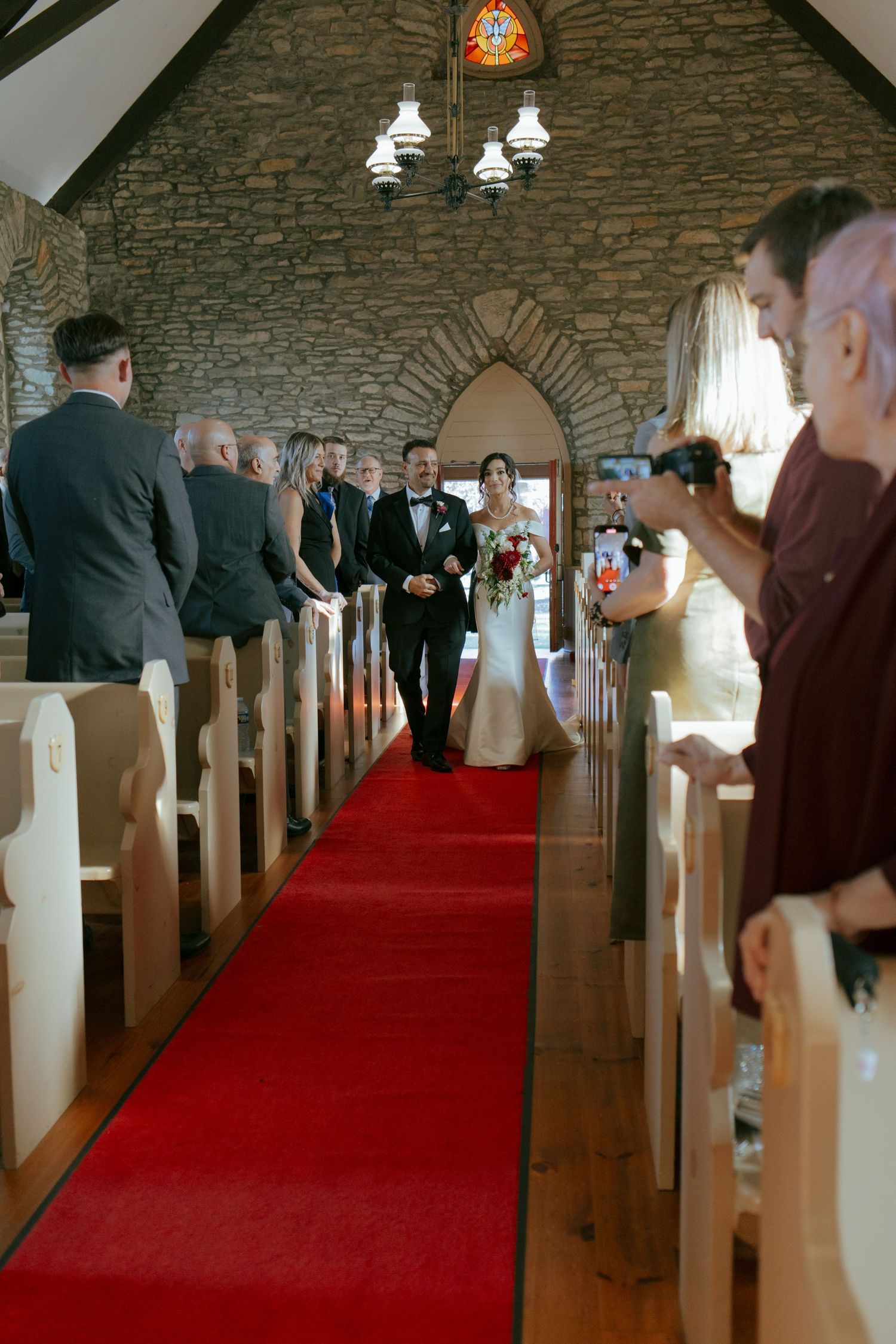 Bride walks down red carpet aisle with escort in stone chapel wedding ceremony.