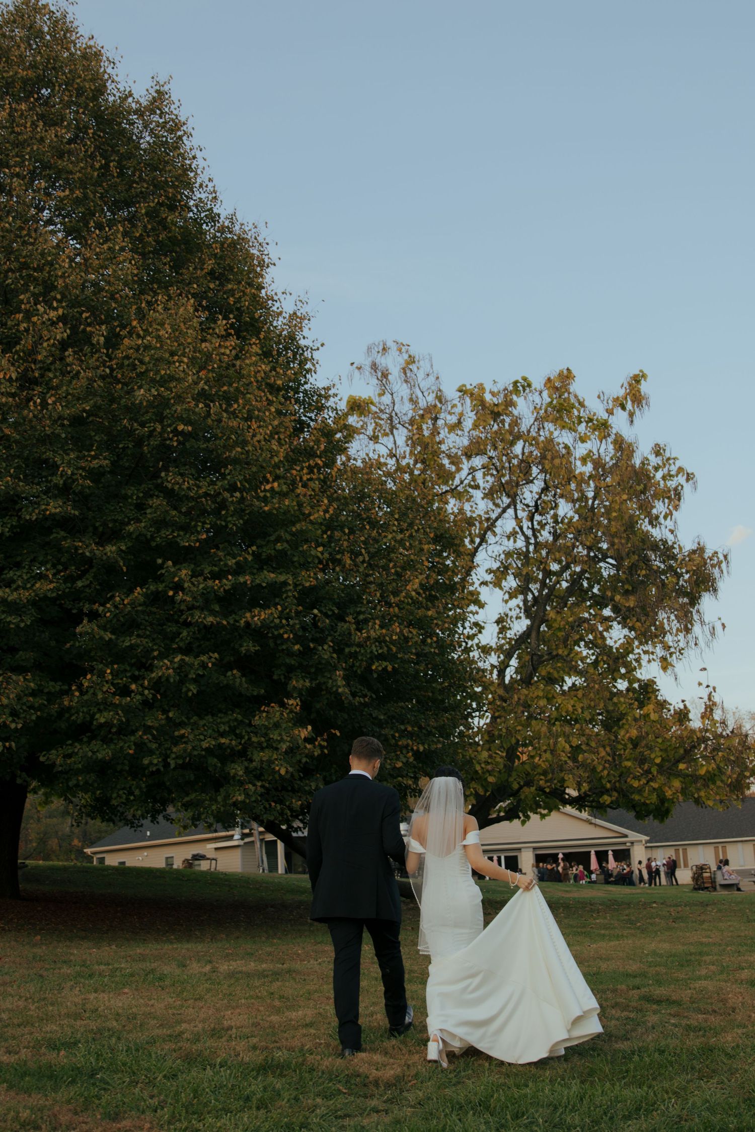 Bride and groom walking hand in hand across green lawn toward trees at outdoor wedding venue during golden hour.