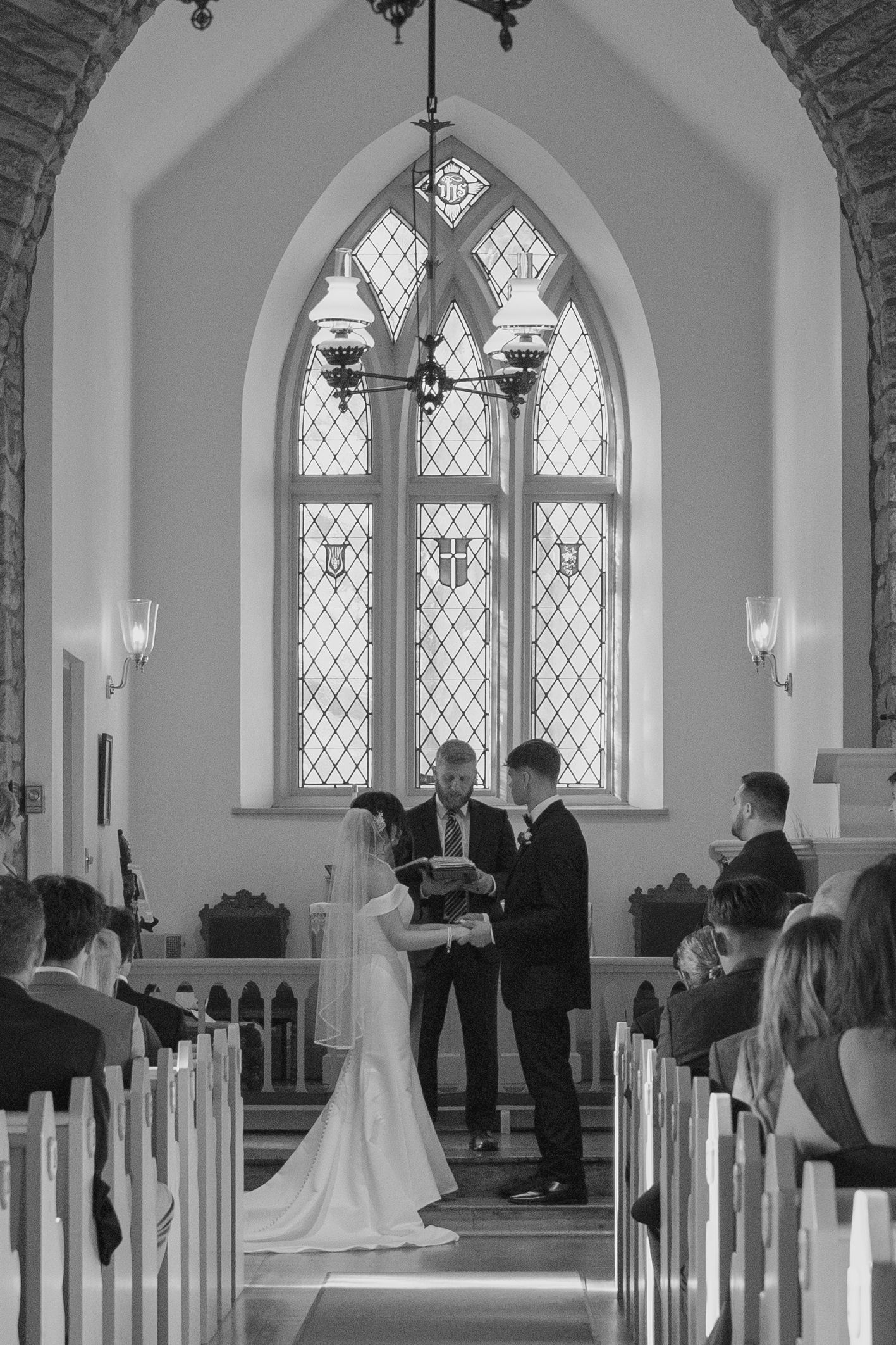 Black and white photo of a bride and groom exchanging vows at a church altar with a stunning arched stained glass window.