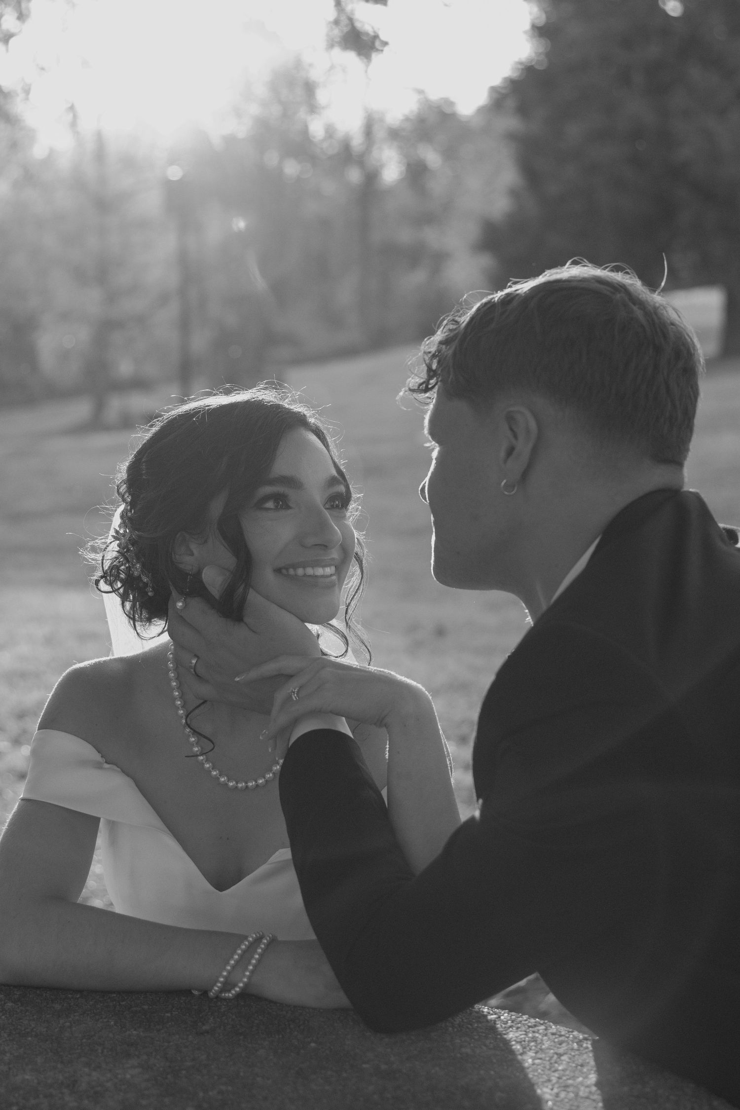 Black and white portrait of a smiling bride wearing pearls as groom tenderly holds her face outdoors.