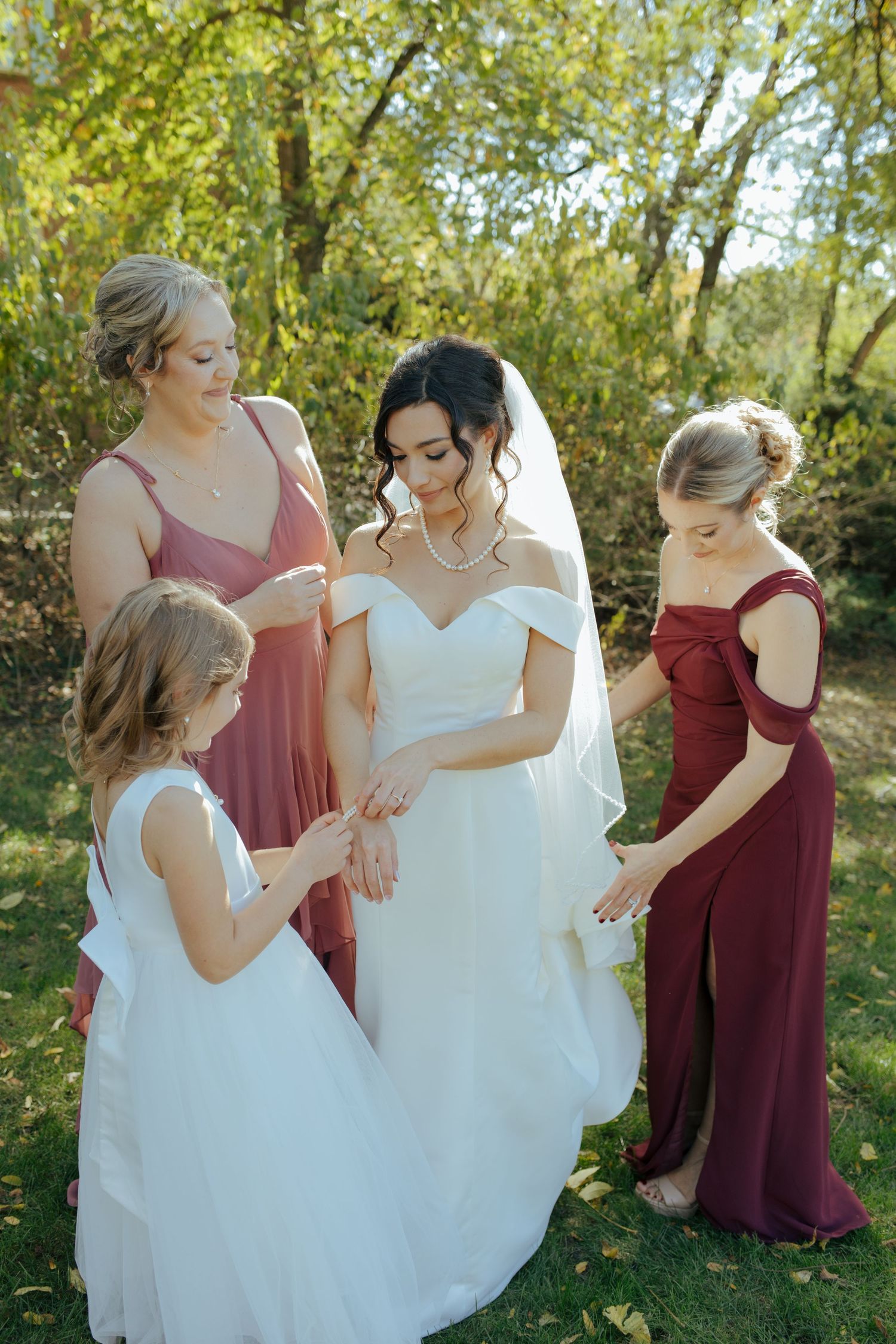 Bride in white gown with bridesmaids in burgundy and mauve dresses outdoors in garden.