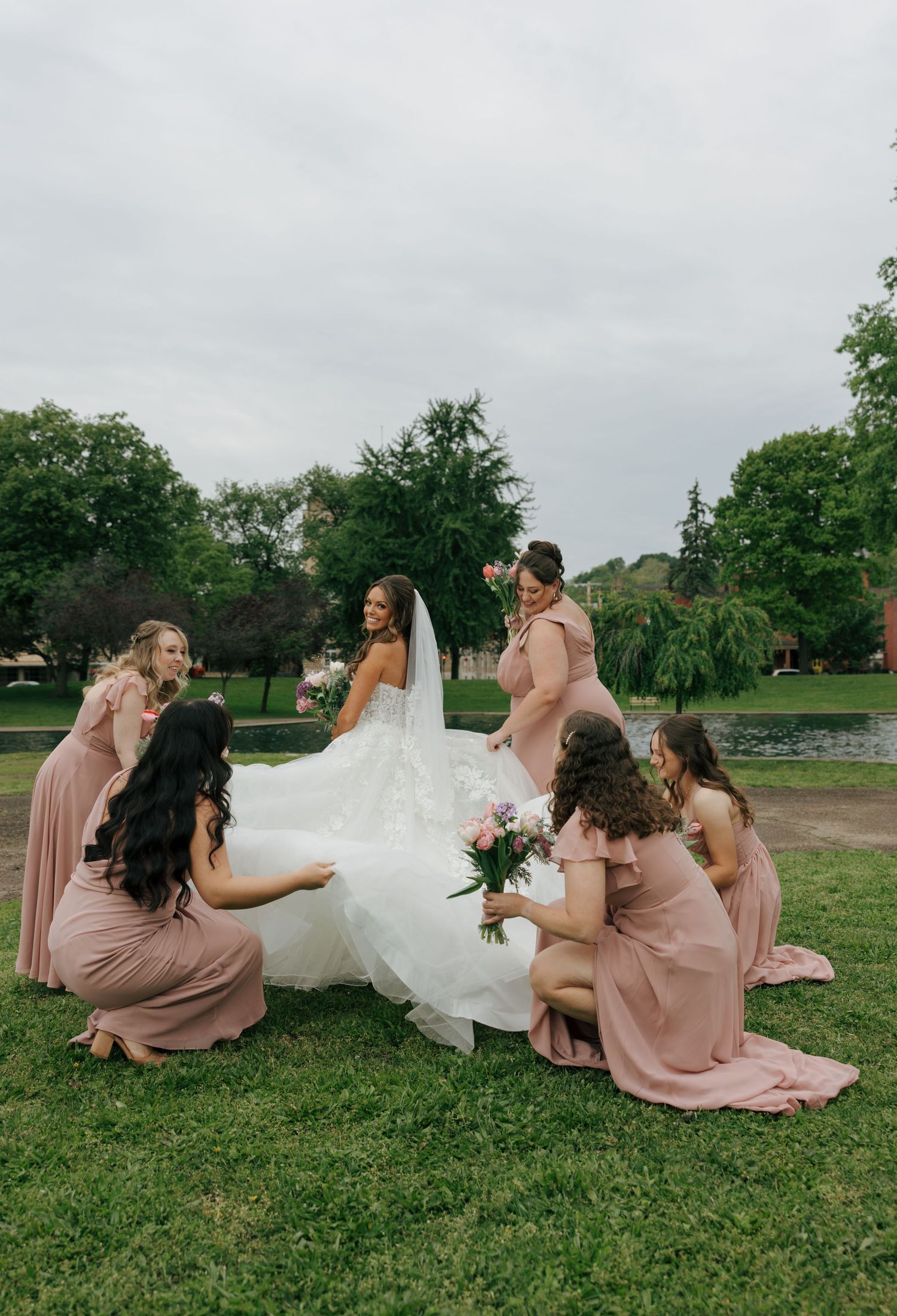 Bride in white gown surrounded by bridesmaids in pink dresses on green lawn near a pond.