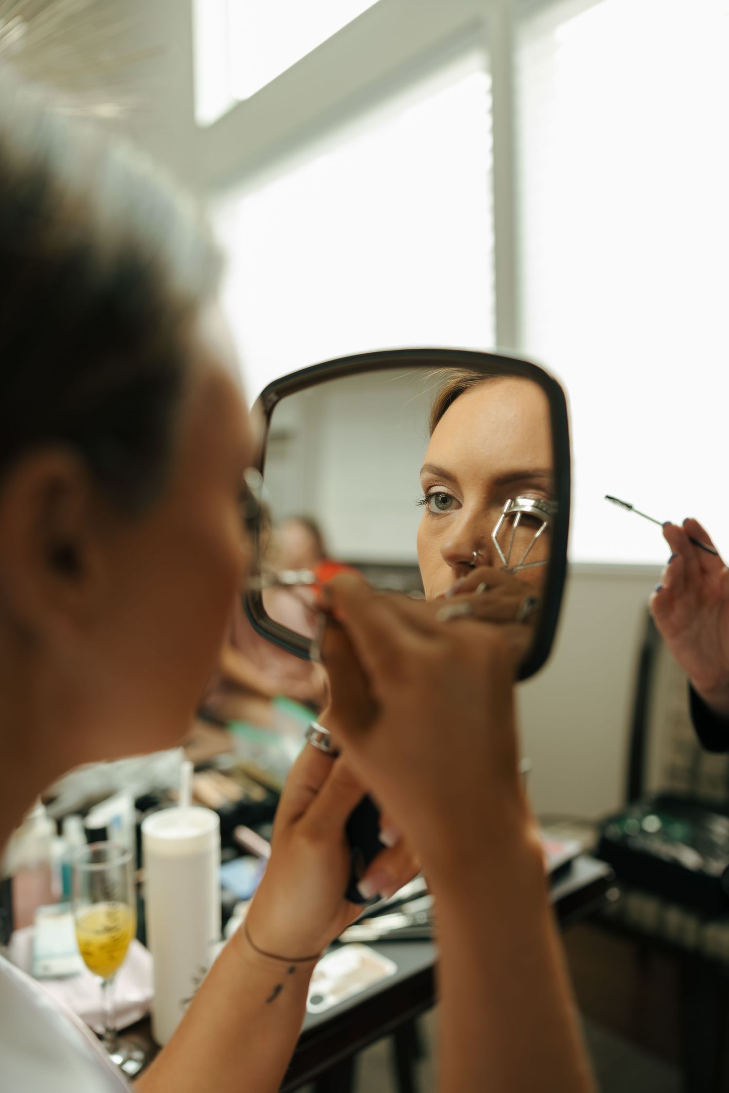 Woman applying mascara while looking at her reflection in a handheld mirror at a makeup vanity.