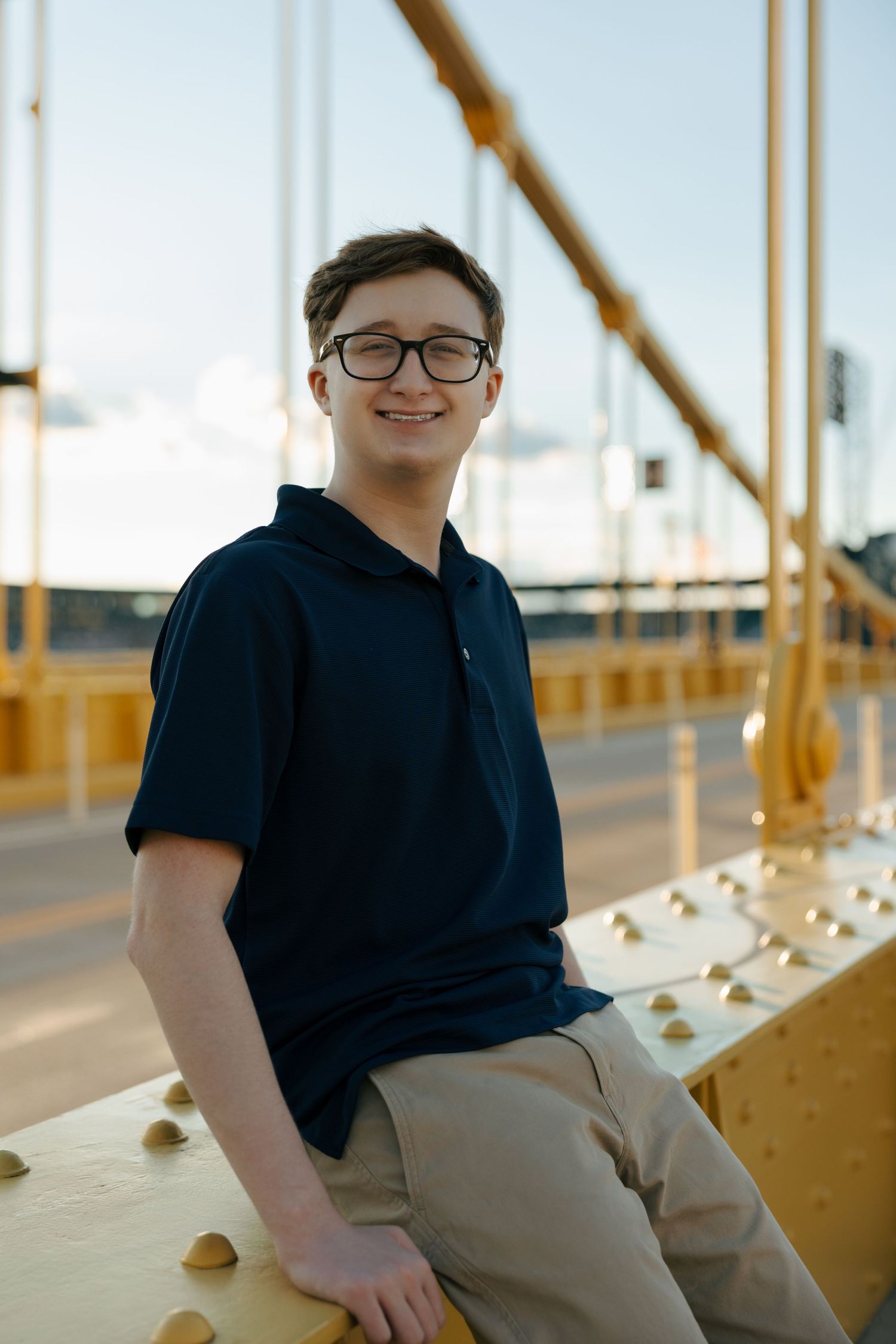 Smiling teen in glasses and navy polo sits on yellow bridge railing at sunset.