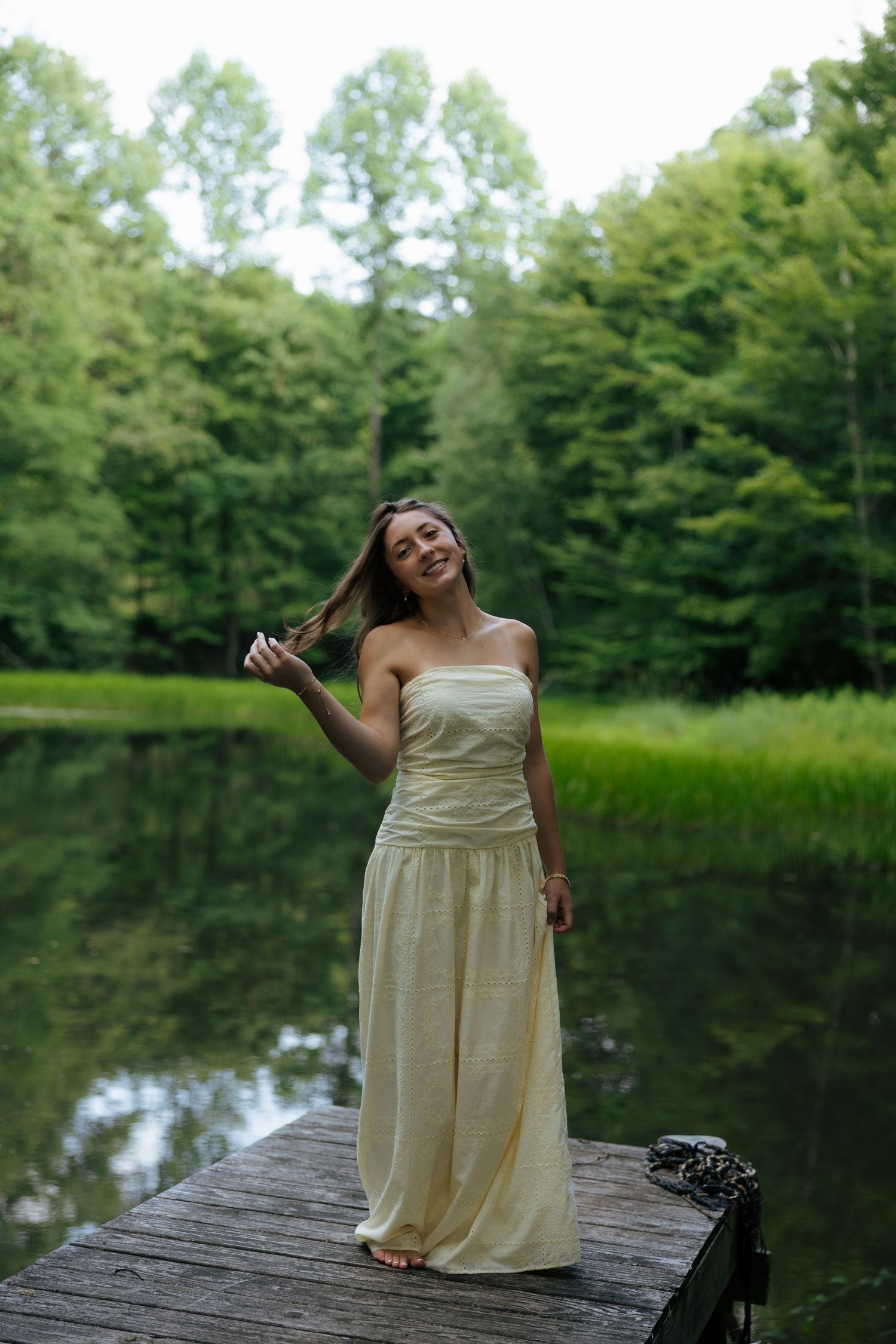 Young woman in cream strapless maxi dress standing on wooden dock by serene forest lake.