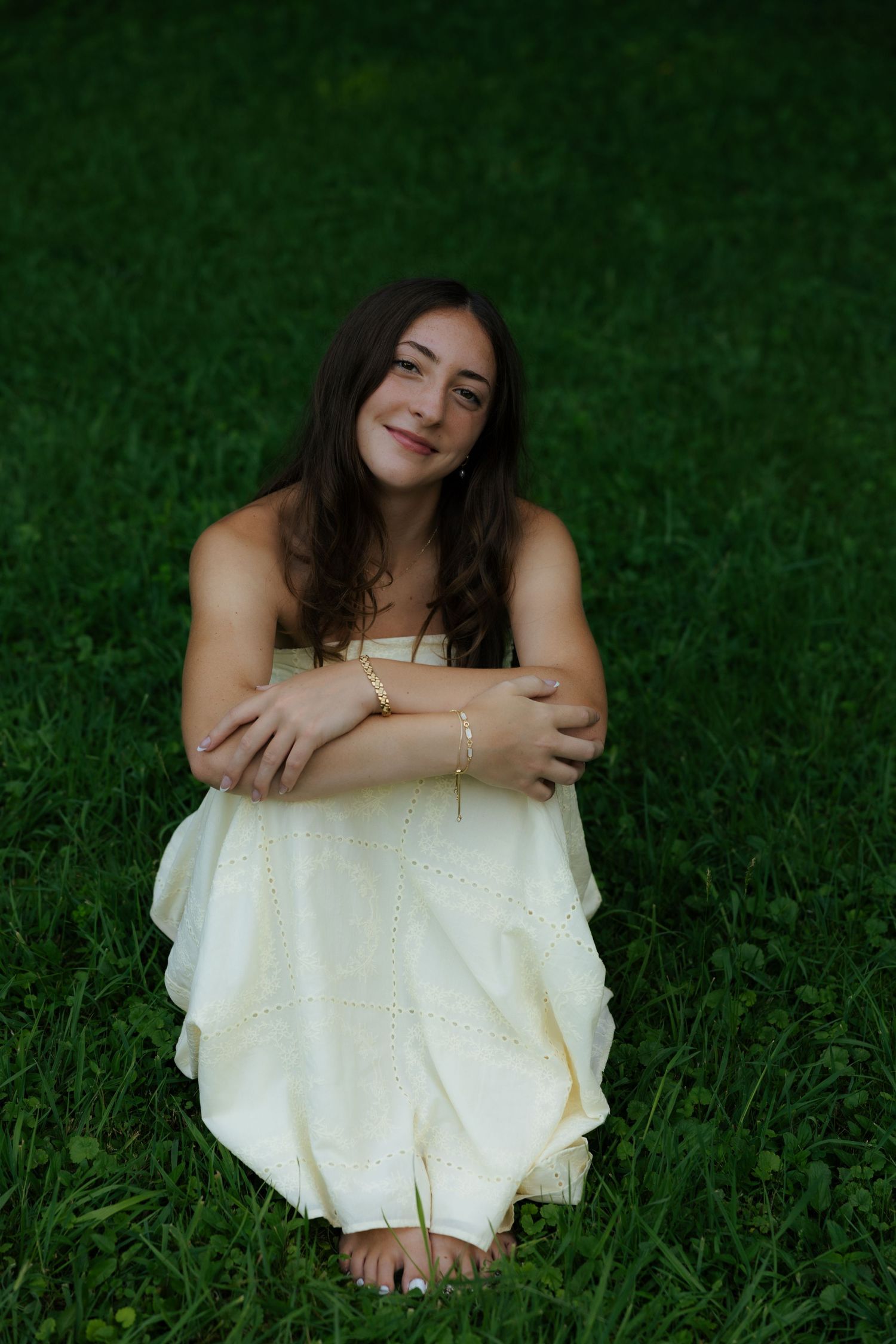 Young woman in a white eyelet dress sitting barefoot on green grass, smiling softly with arms wrapped around knees.