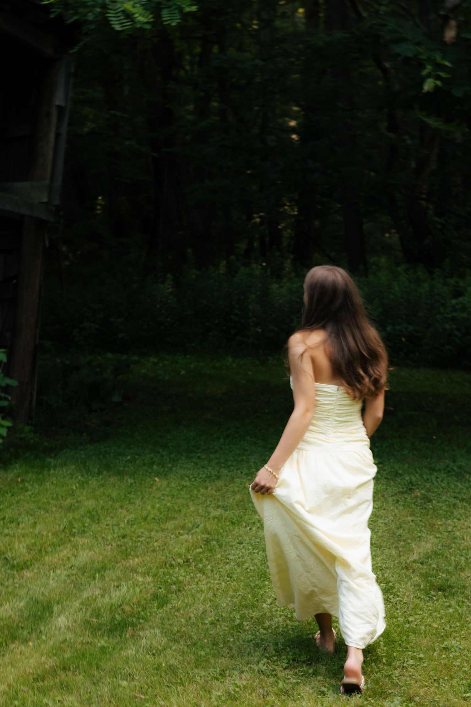 Young woman in flowing yellow strapless gown walking barefoot across green lawn toward dark forest.