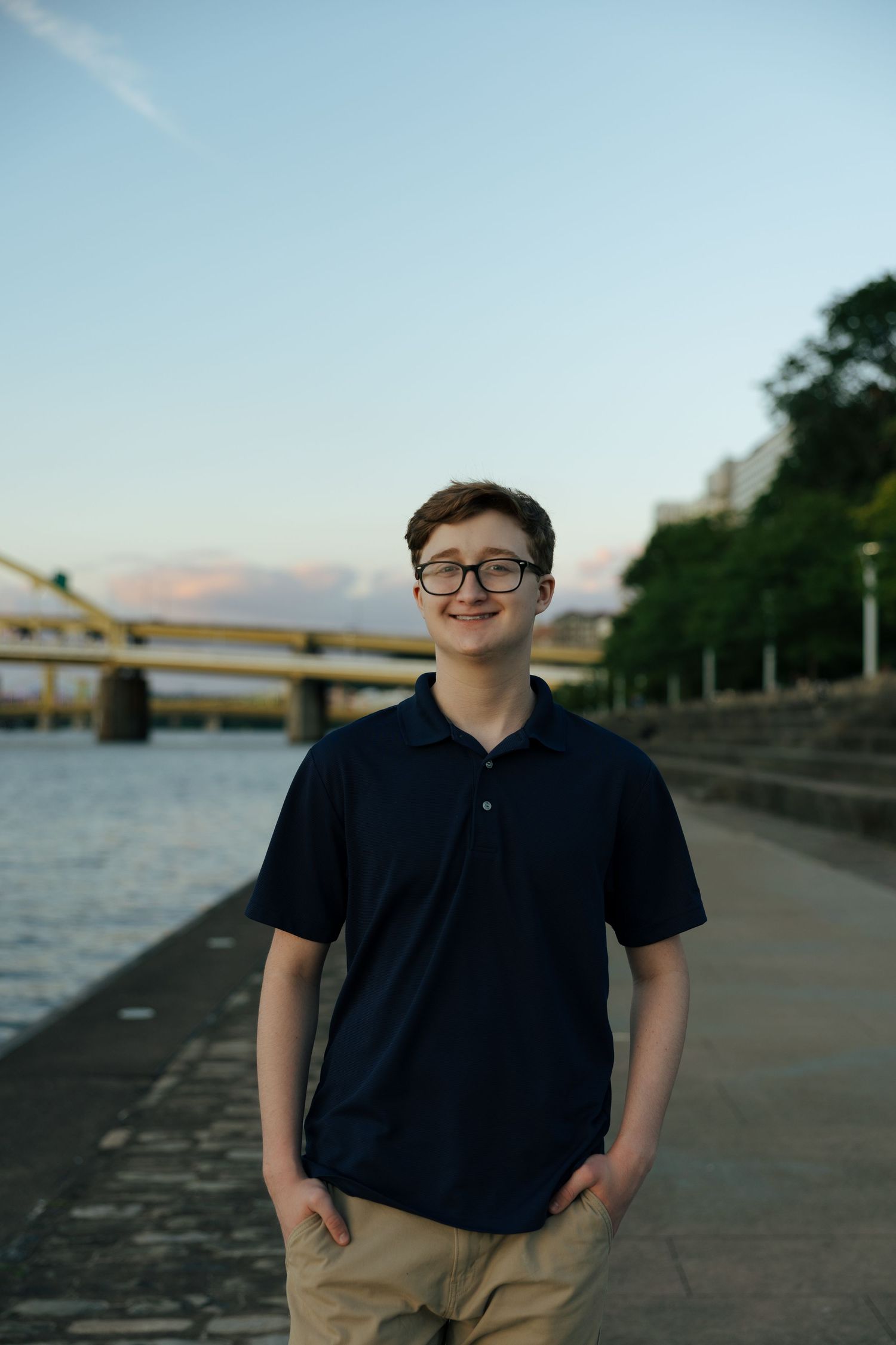 Smiling teen boy in navy polo standing on a Pittsburgh riverfront walkway at dusk.