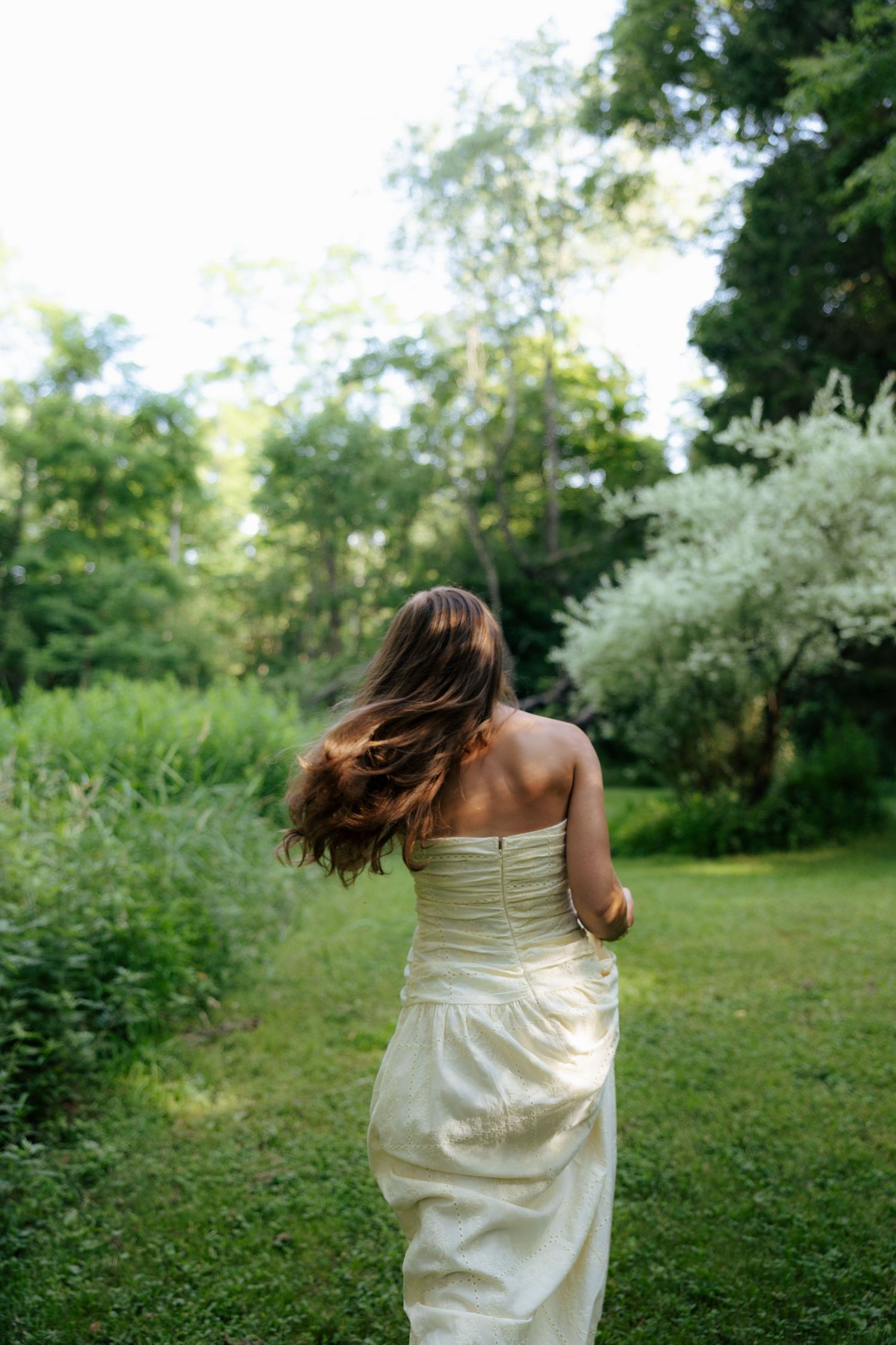 Young woman in strapless cream gown walking away through lush green garden path.