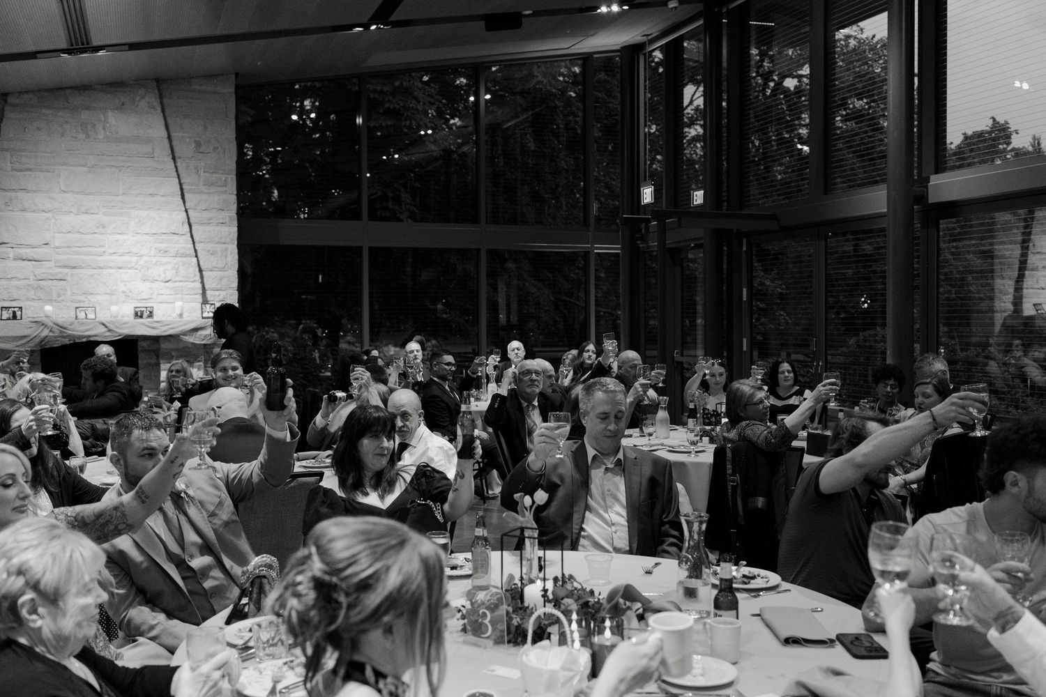Black and white photo of guests raising glasses at an elegant indoor dinner event with large windows.
