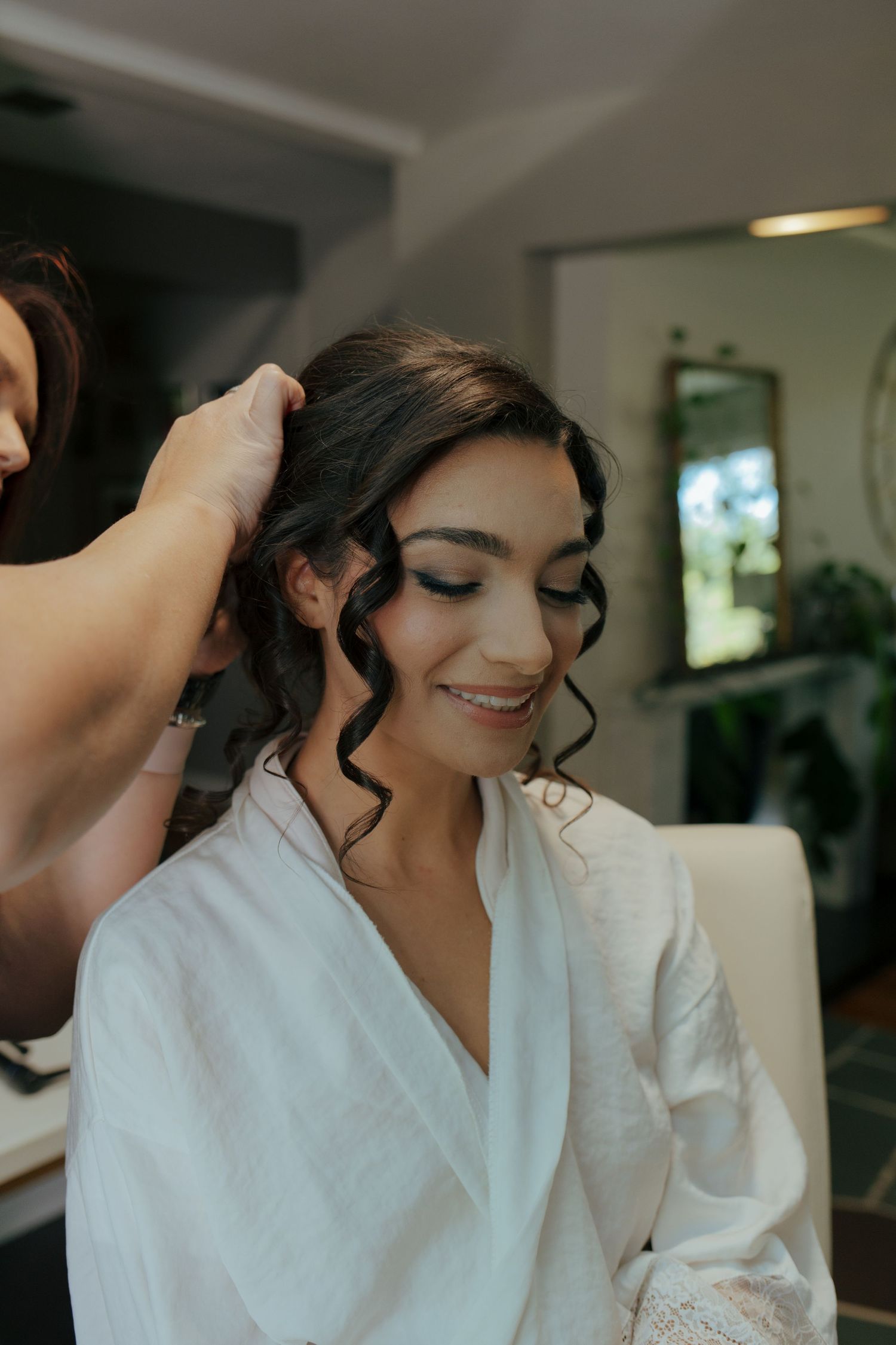 Smiling bride in white robe having her dark curly hair styled by a hairdresser.