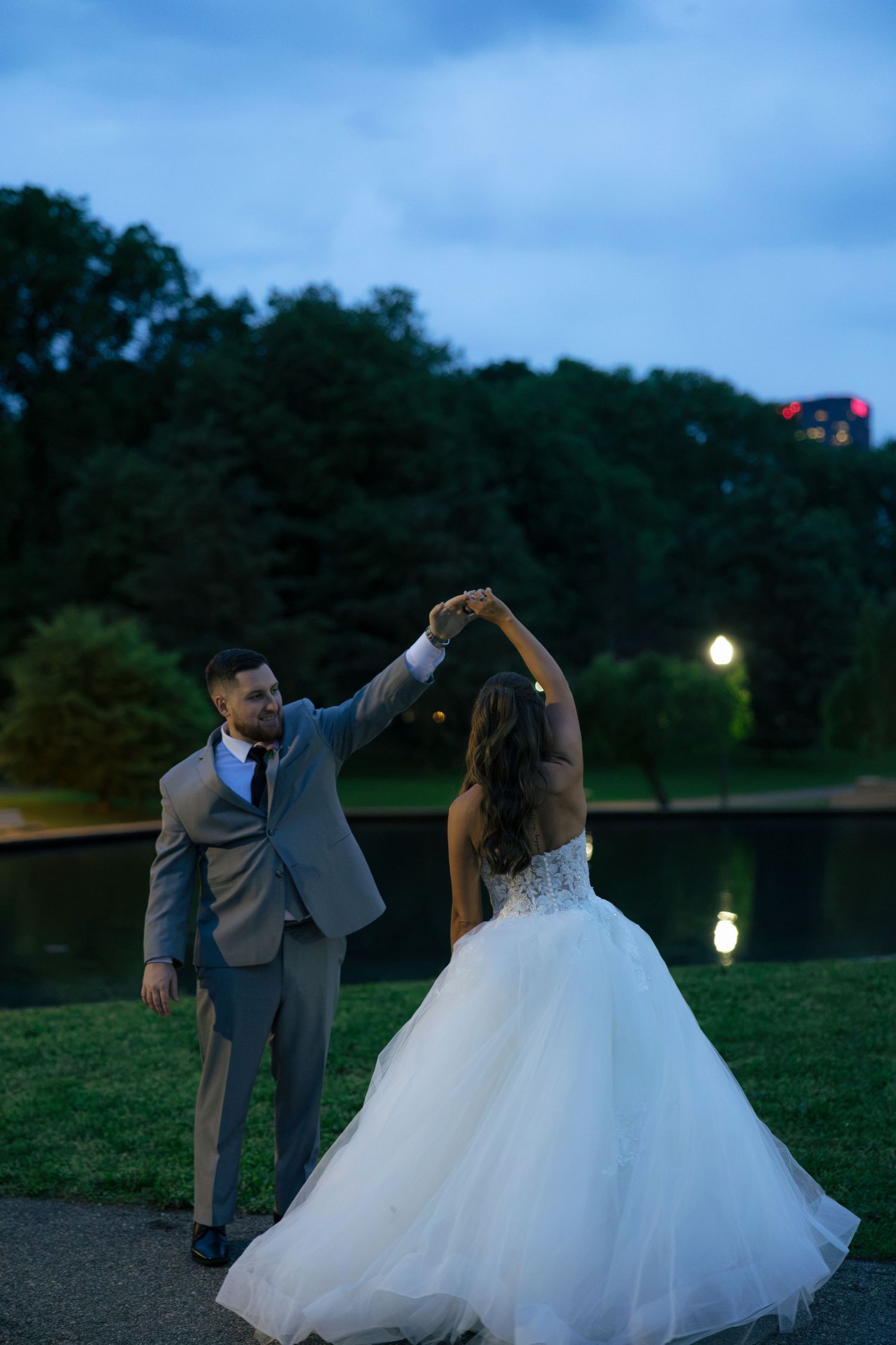 Bride and groom dancing outdoors at dusk near a pond, groom in gray suit twirling bride in white ballgown.