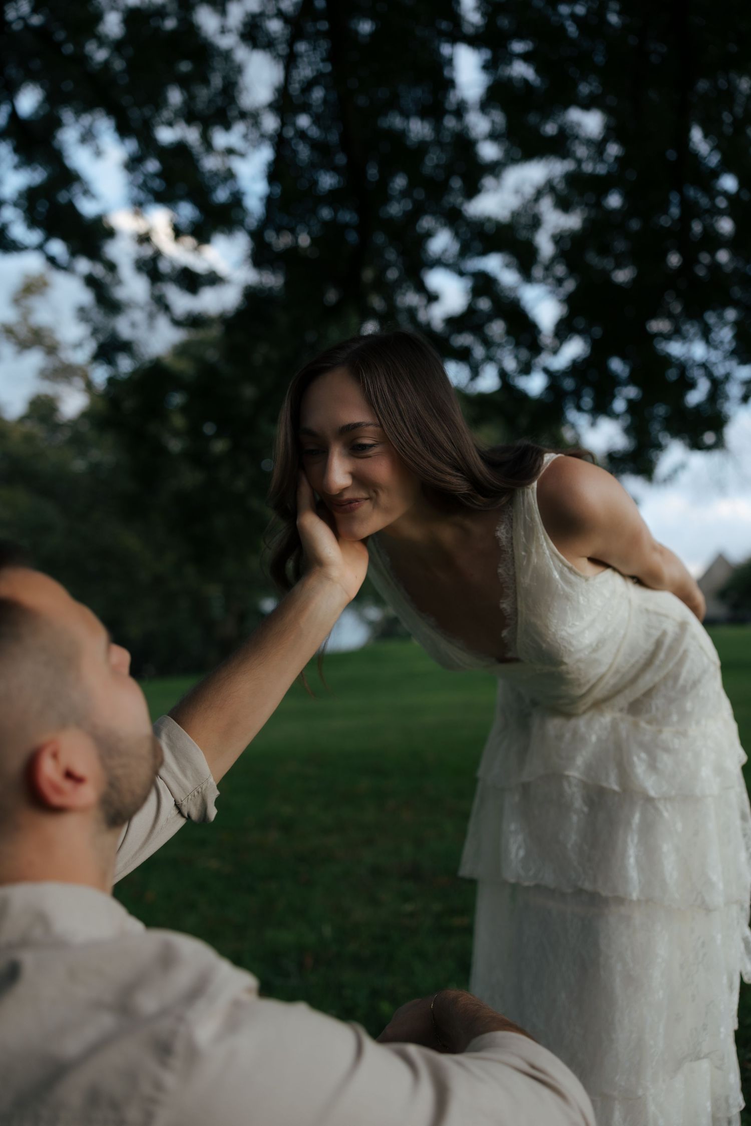 Groom tenderly touches brides cheek as she leans toward him in lush green park.