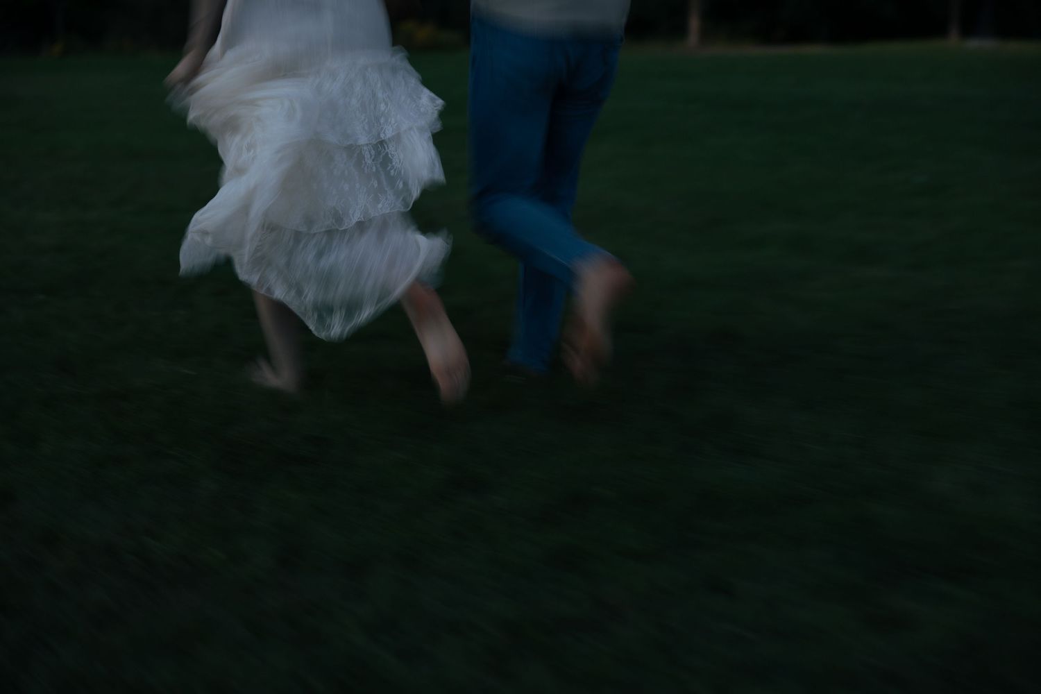 Blurred motion shot of a couple running barefoot on grass at dusk in wedding attire.