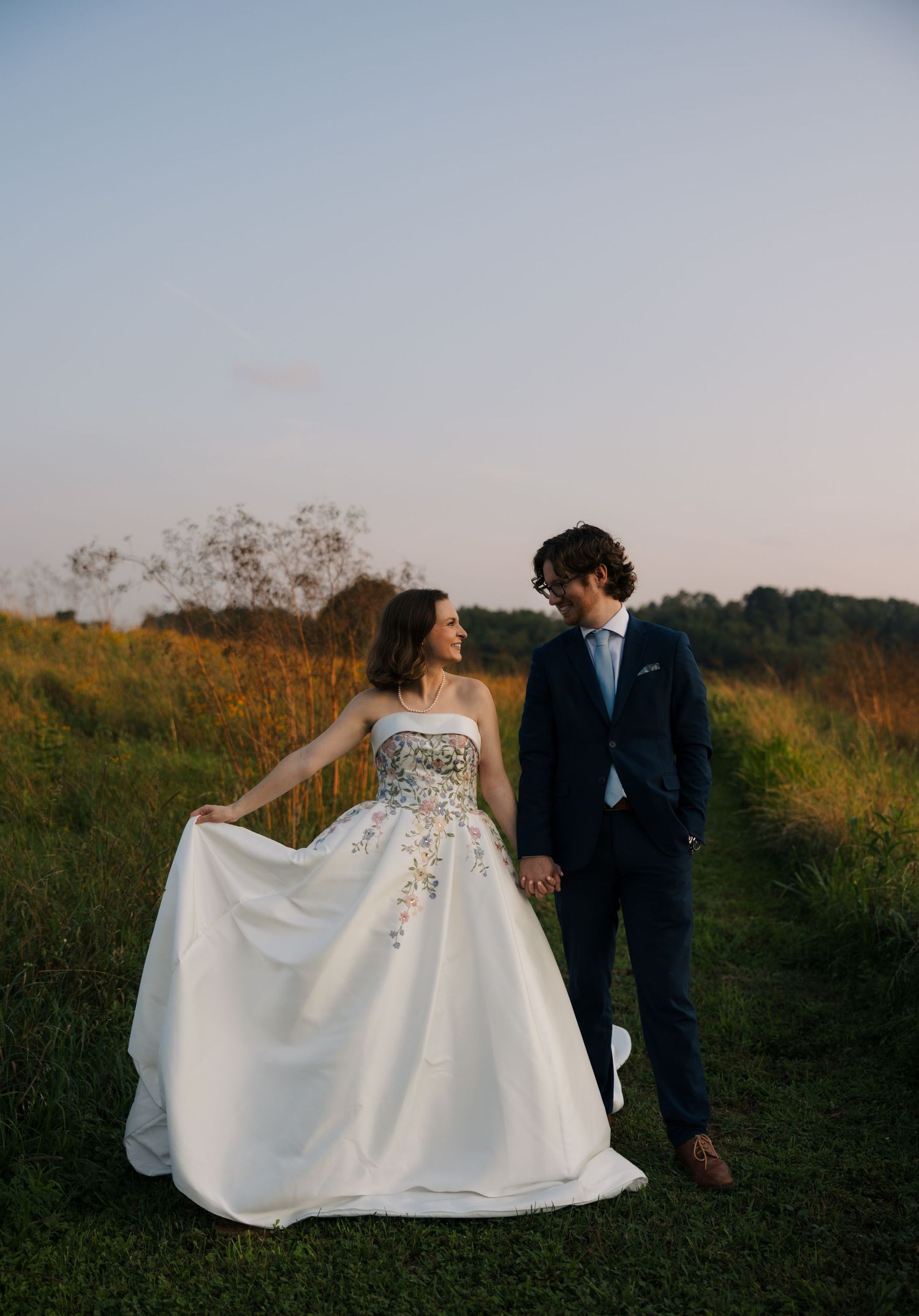 Bride in floral strapless gown holds groom's hand in navy suit walking through golden meadow at sunset.