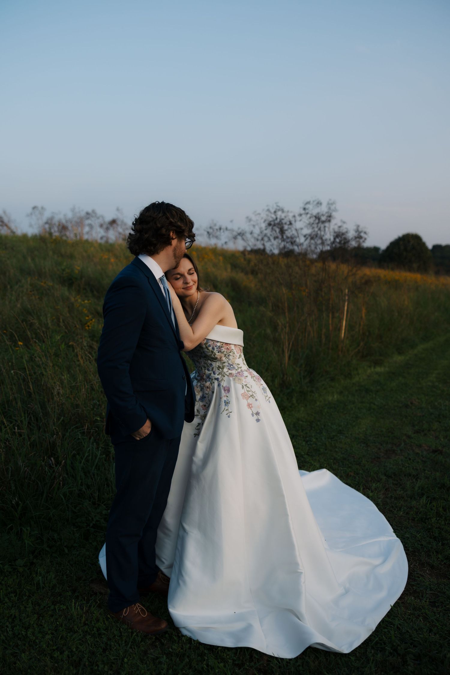 Bride in floral strapless gown leans on groom in navy suit during golden hour outdoor wedding portrait.
