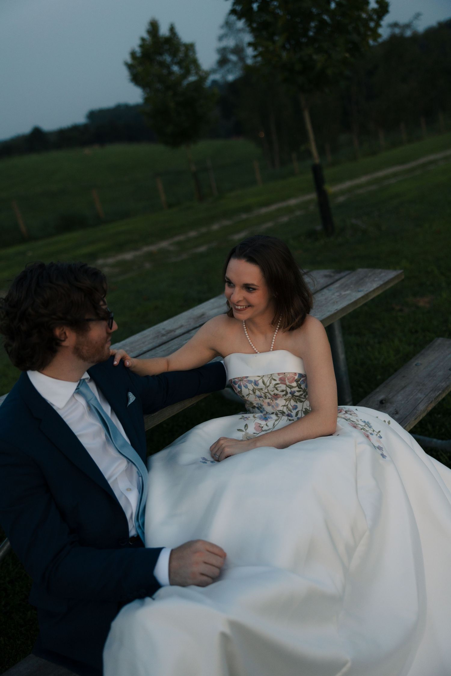 Bride and groom sitting on wooden bench at dusk, smiling at each other in outdoor farm setting.
