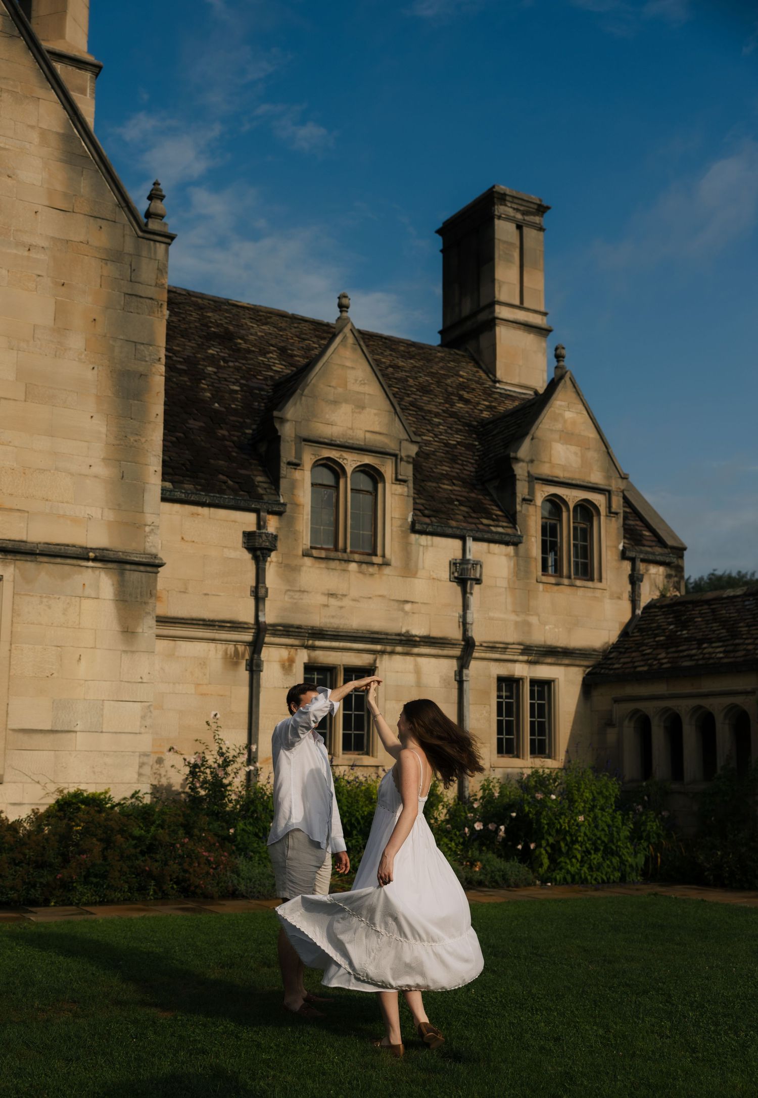 Couple dancing joyfully on green lawn before a grand stone manor under blue sky.