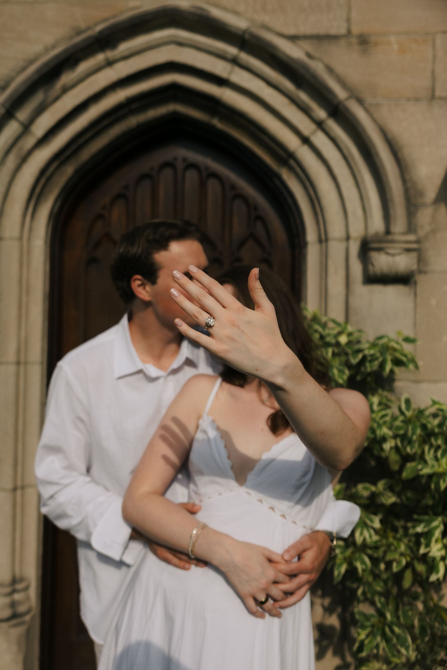 Couple embracing under a Gothic stone archway, woman showing off her diamond engagement ring.