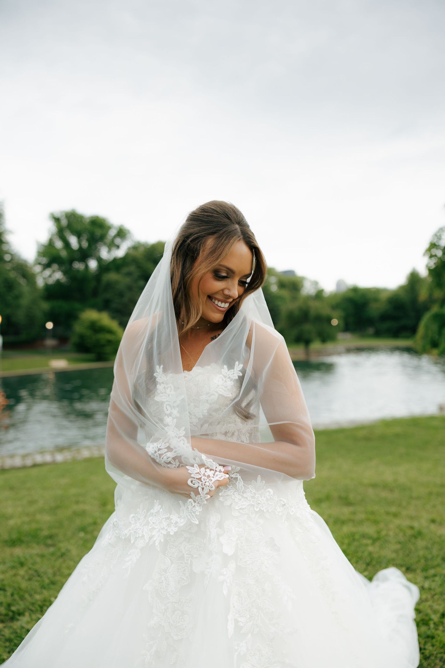 Smiling bride in white lace wedding gown and veil poses outdoors near a pond surrounded by green trees.
