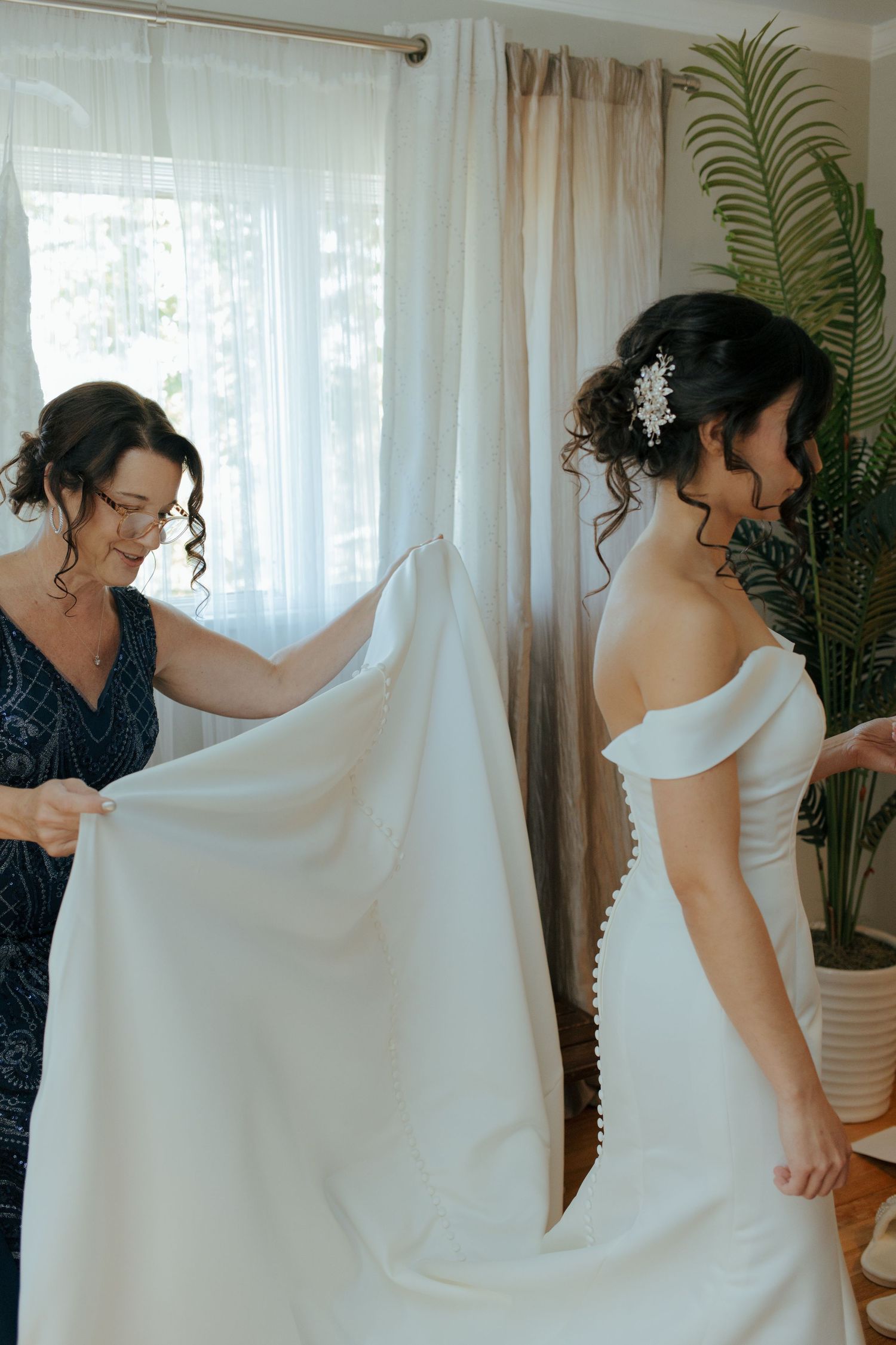 Bride in white off-shoulder gown gets help with train from bridesmaid in navy dress indoors.