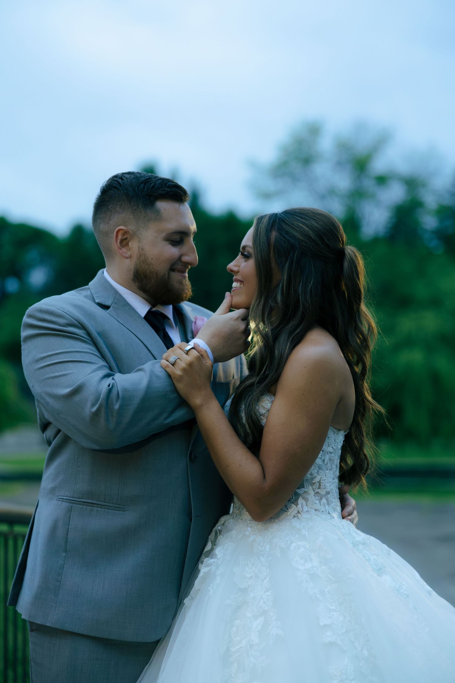 Smiling bride in a lace wedding gown laughs joyfully as groom in gray suit tenderly touches her face at twilight.