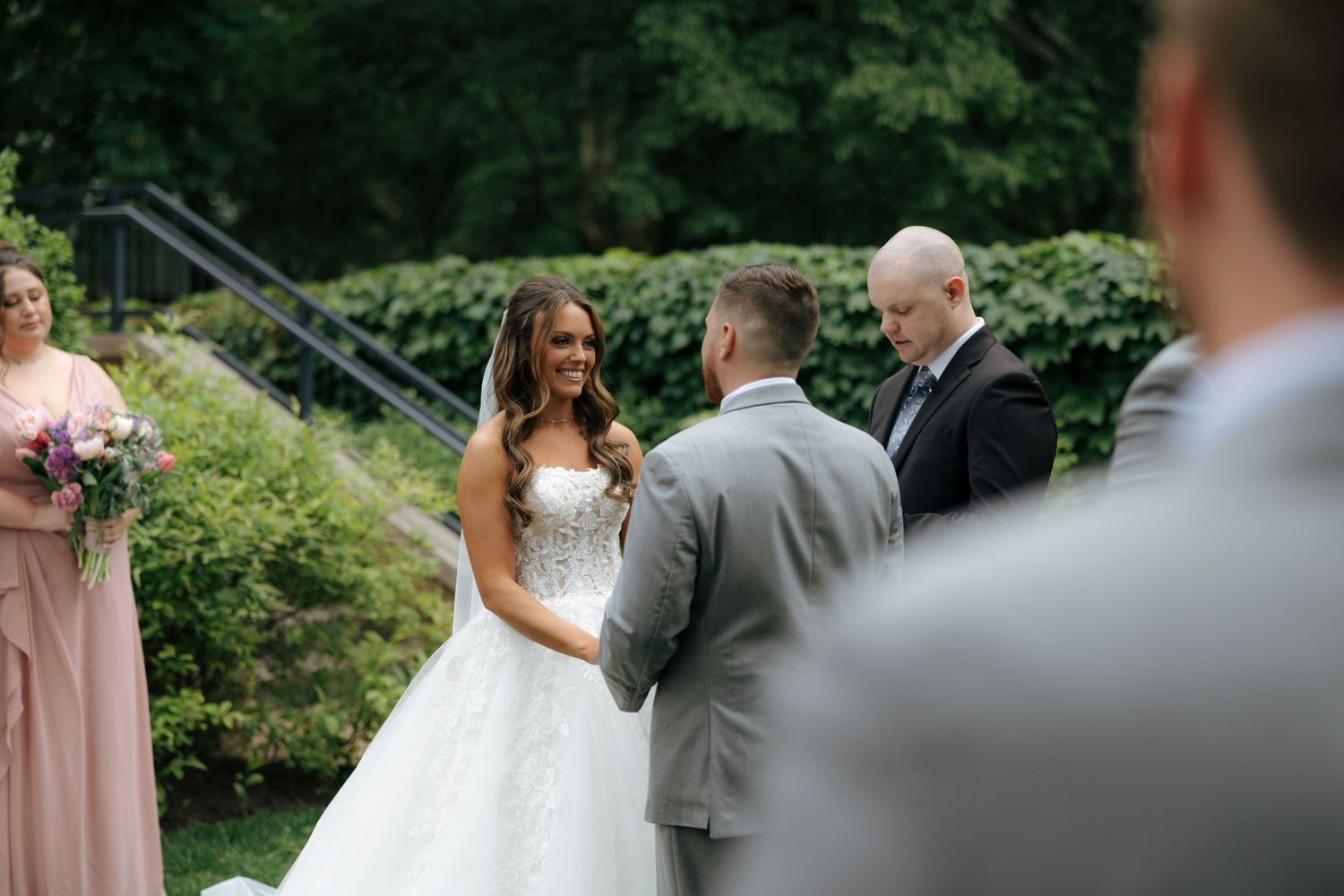 Smiling bride in lace ballgown holds hands with groom in gray suit during outdoor wedding ceremony.