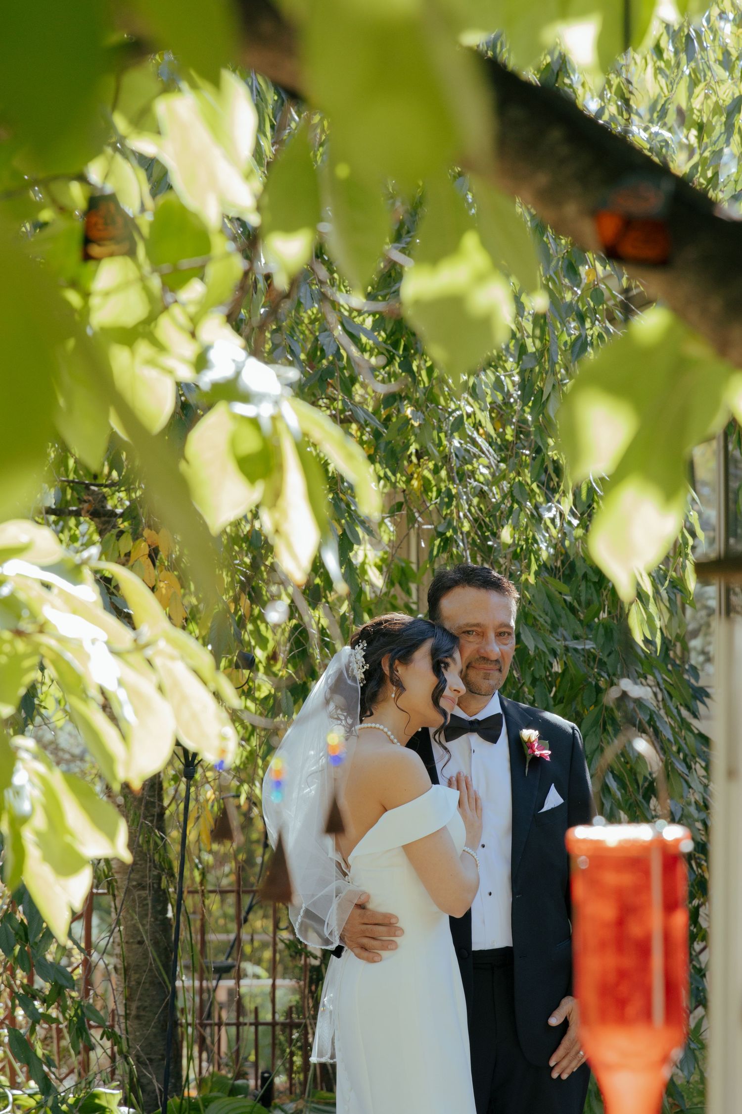 Bride and groom share a romantic moment in a lush garden, framed by green leaves, as she touches his chest.