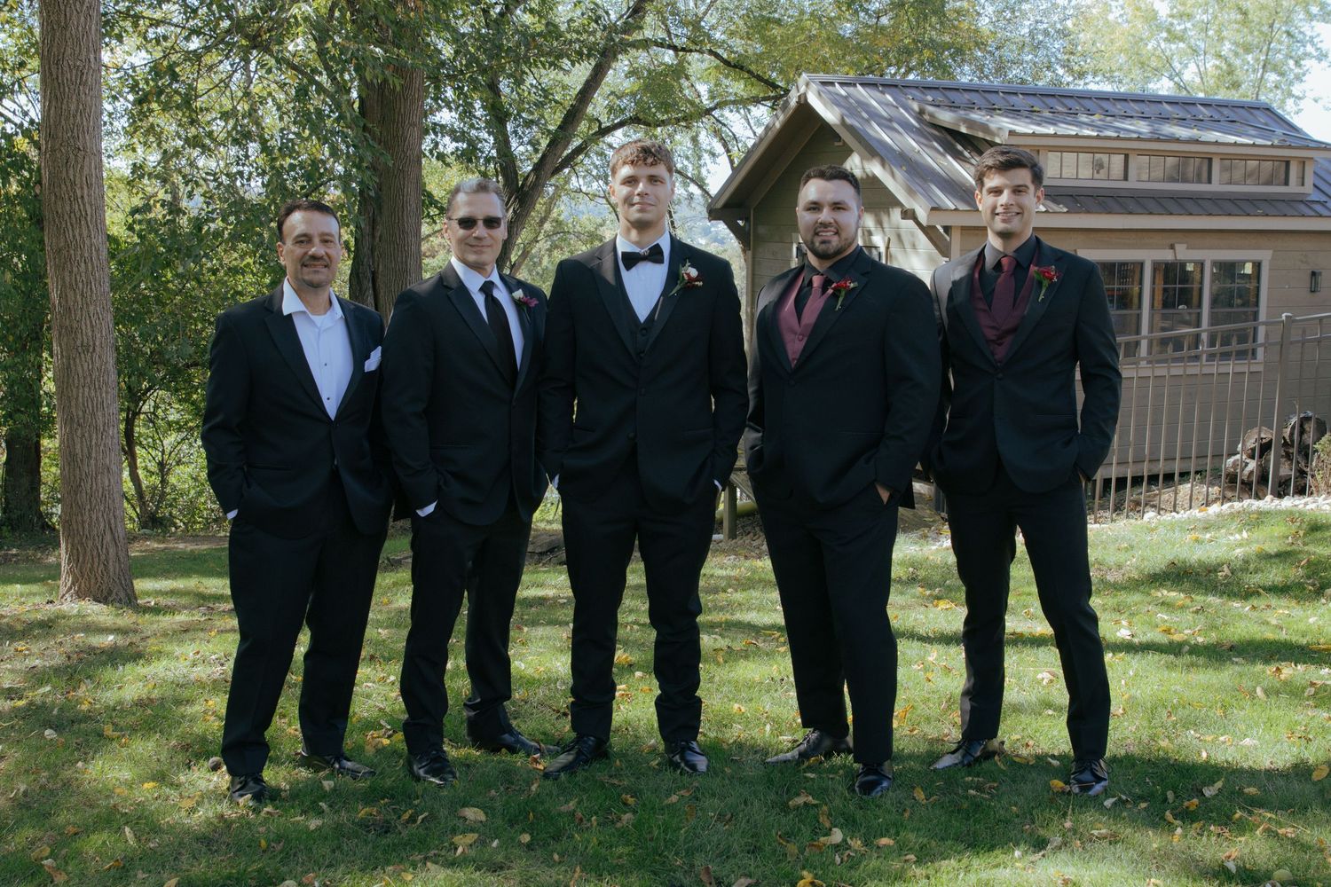 Five groomsmen in black tuxedos pose outdoors near a rustic cabin for a wedding photo.