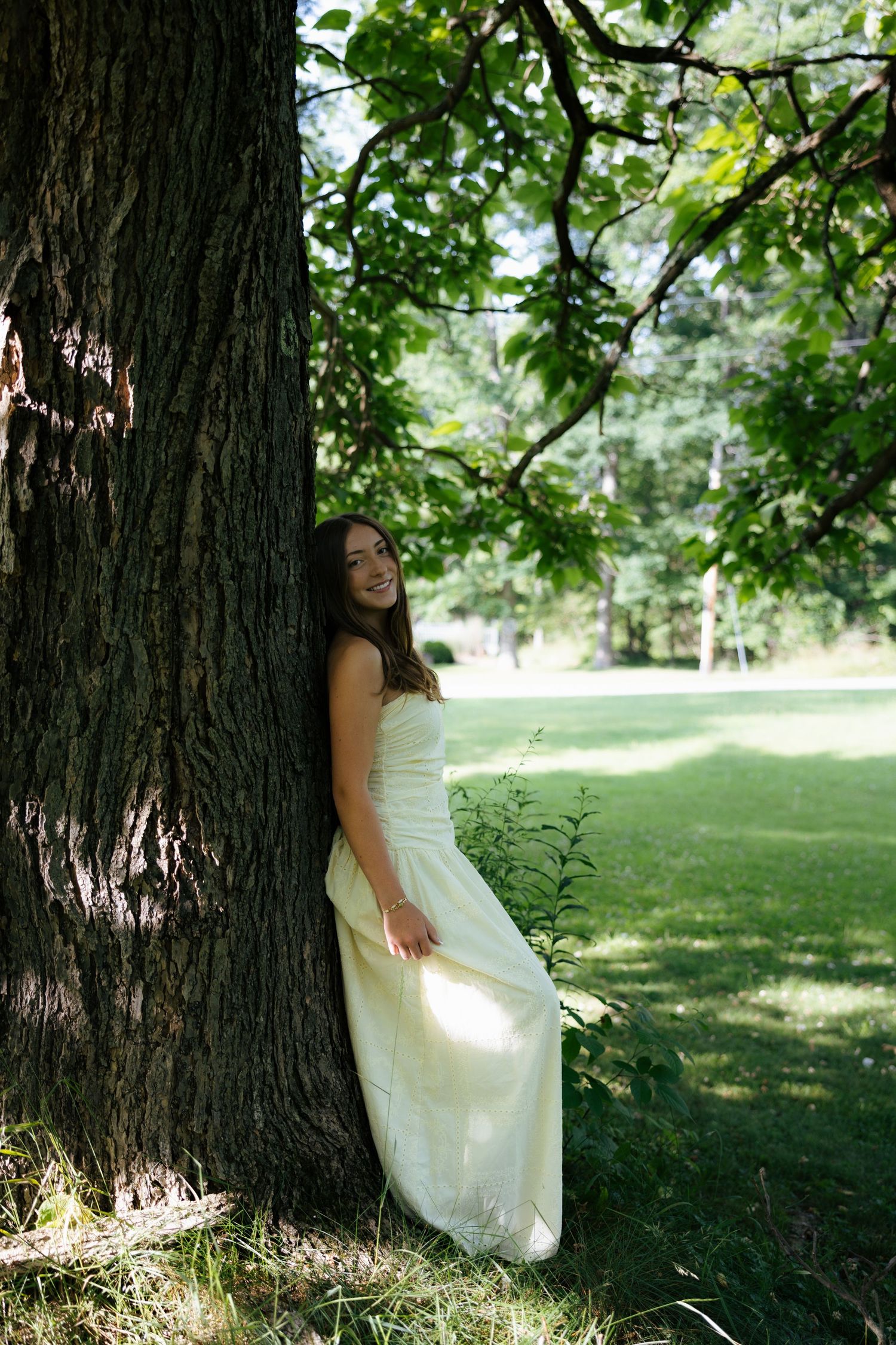 Smiling young woman in elegant cream strapless gown leaning against large tree in sunny park.