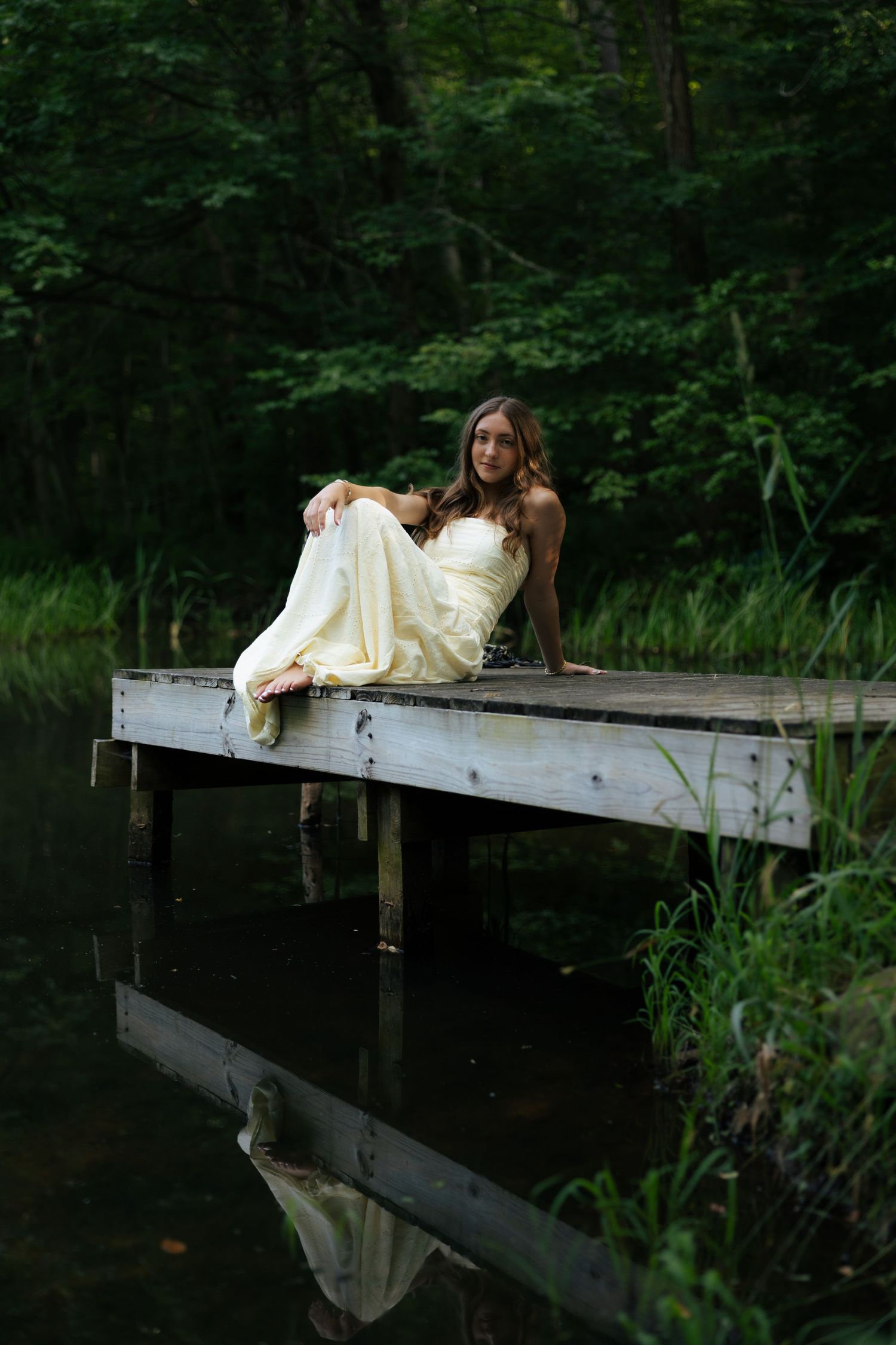 Young woman in yellow strapless gown sitting on wooden dock reflected in calm forest pond.