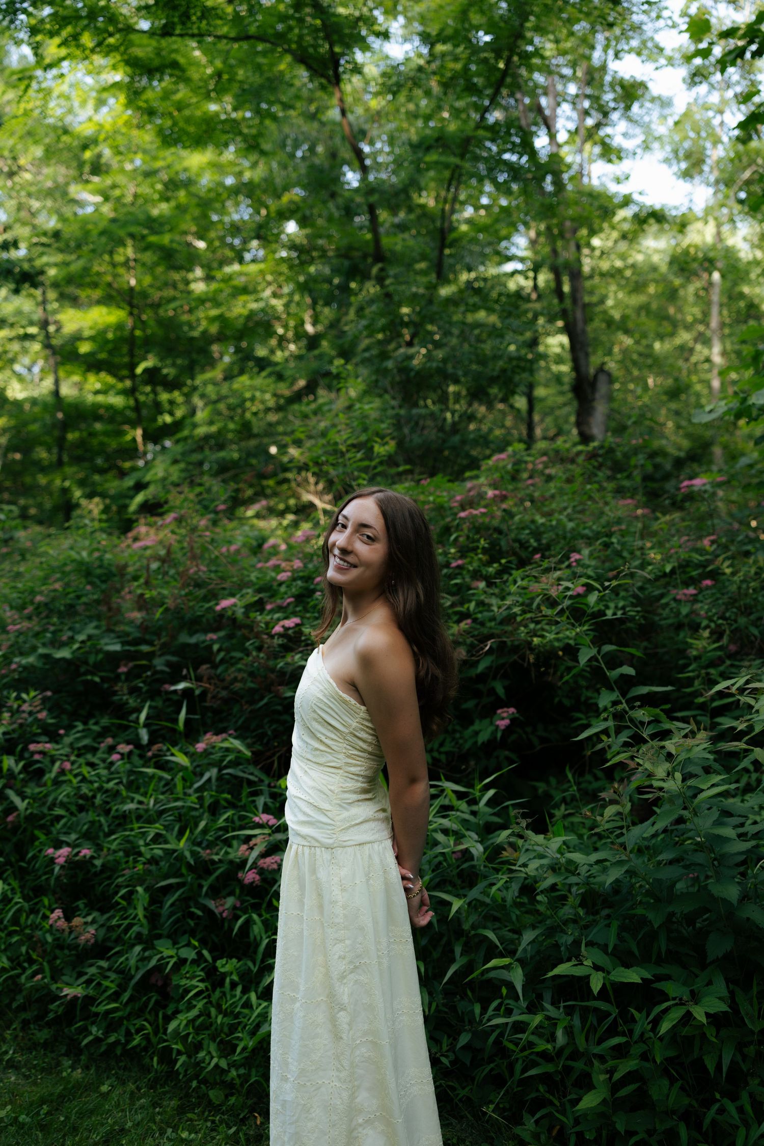 Smiling young woman in white strapless gown standing among pink flowering garden shrubs.