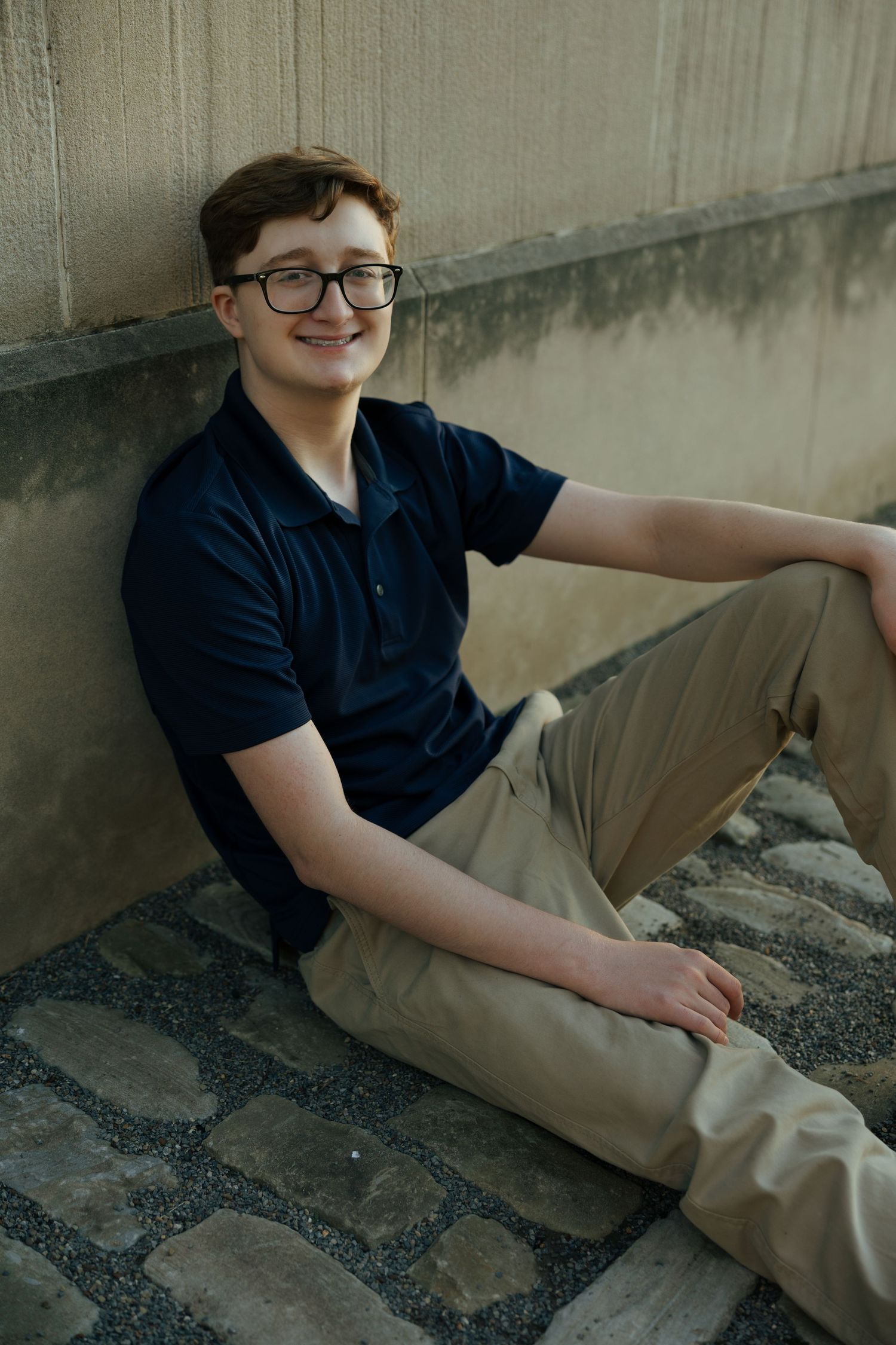 Teen boy in navy polo and khakis sits relaxed against concrete wall on cobblestone ground.