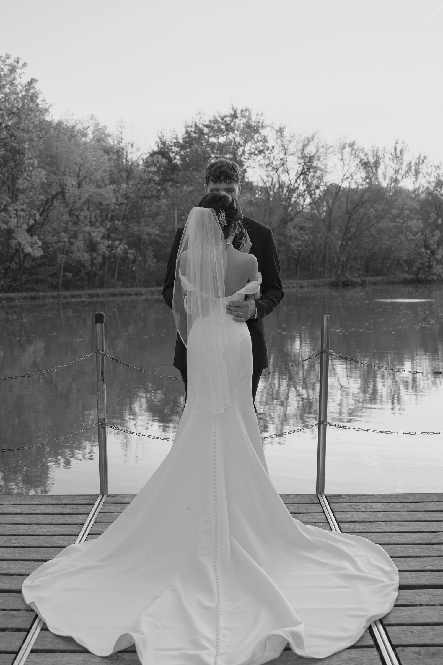 Bride and groom standing on a dock overlooking a calm lake in a black and white wedding photo.