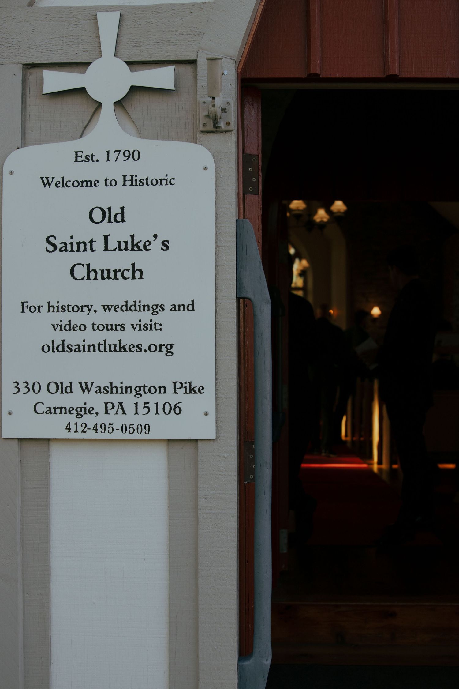 Sign for Historic Old Saint Luke's Church established 1790 in Carnegie PA with open doors glowing warmly inside.