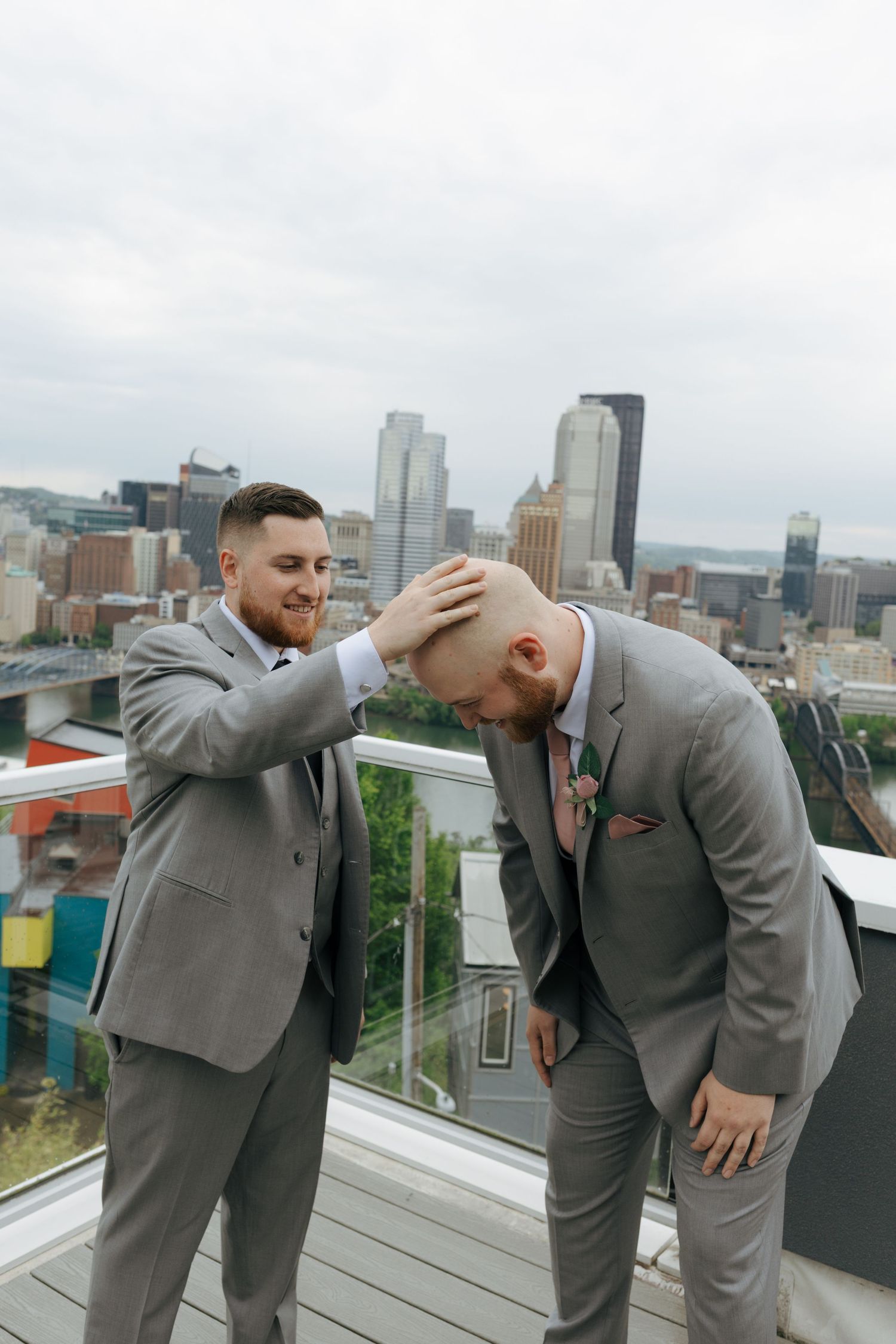 Two groomsmen in gray suits share a lighthearted moment on a rooftop with Pittsburgh city skyline backdrop.