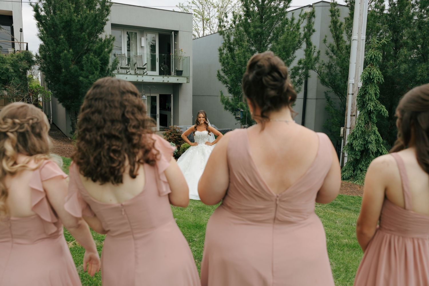 Bridesmaids in blush pink dresses walk toward a bride in a white gown on a green lawn.