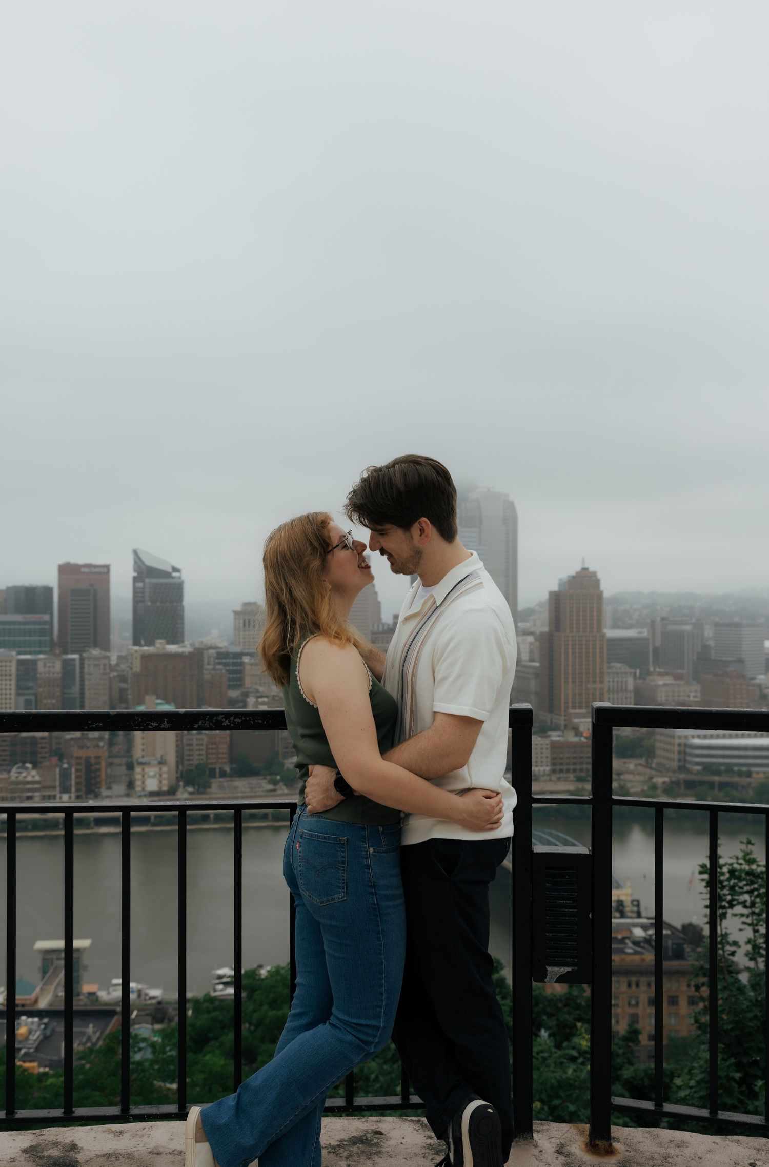 A couple shares an intimate moment nose-to-nose on a rooftop overlook with the Pittsburgh skyline in the background.