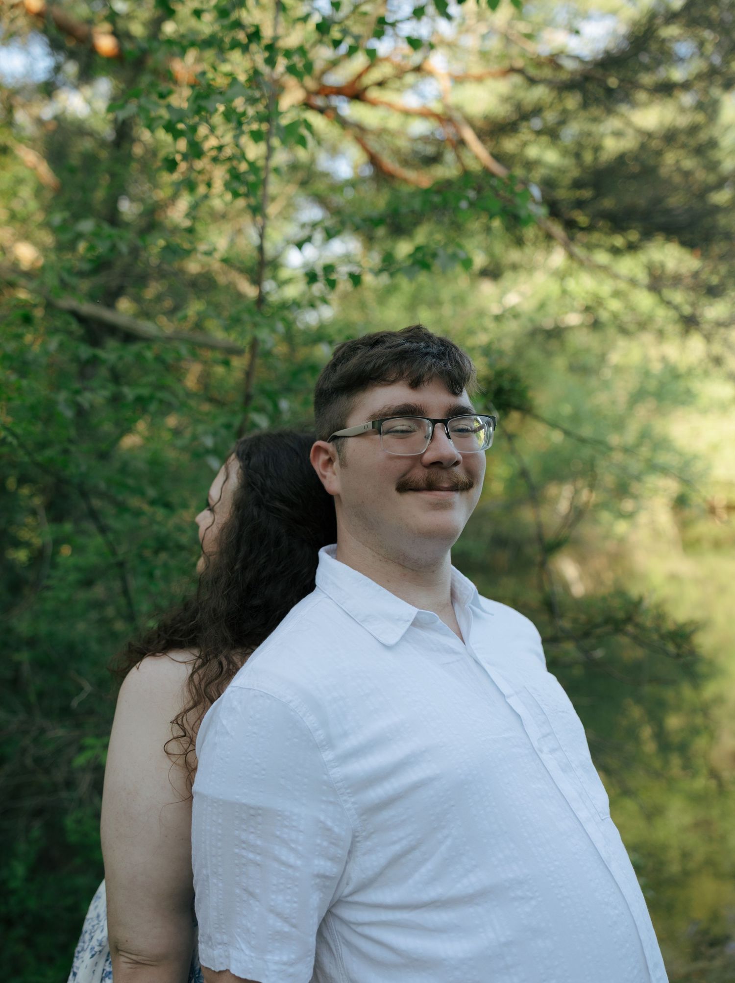 A man with glasses and a mustache in a white shirt stands outdoors with a woman, surrounded by lush green trees.