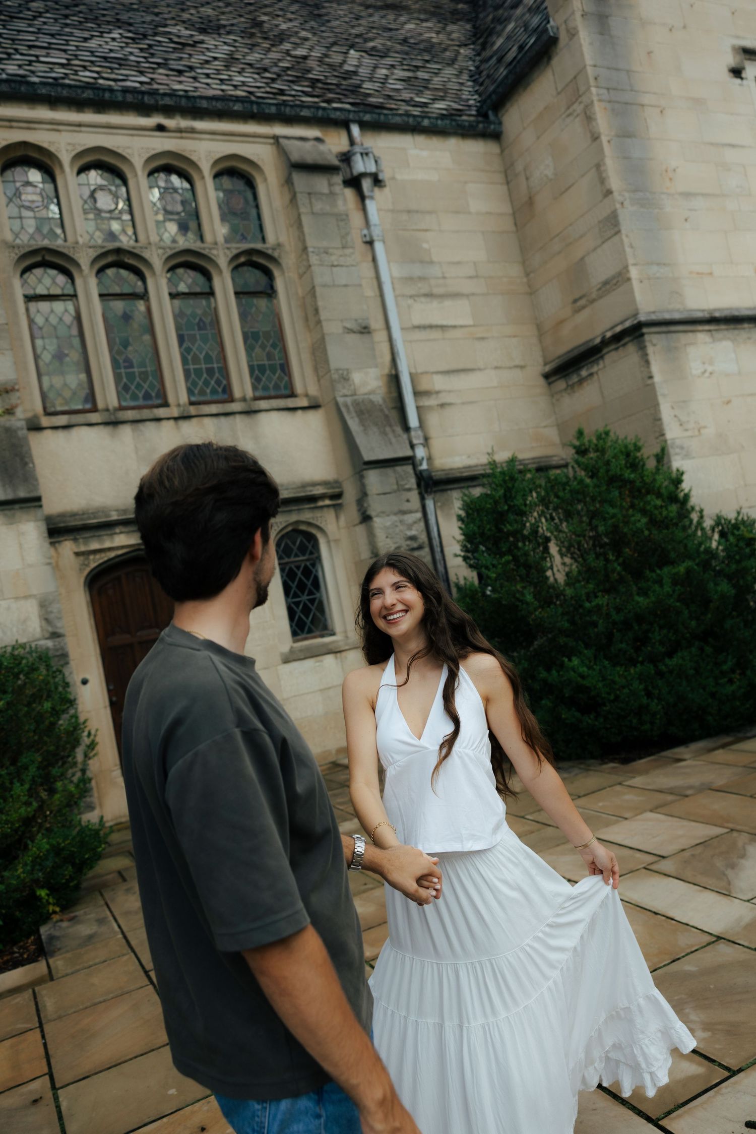 Smiling woman in white dress twirls beside man in gray tee outside a historic stone building.
